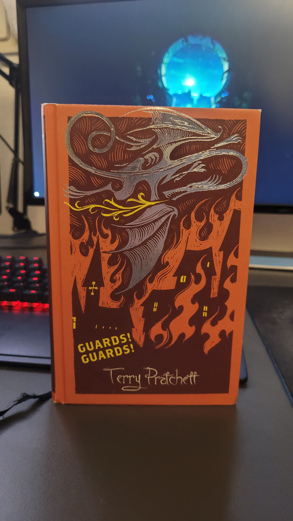 Das Bild zeigt eine Hardcover Ausgabe von Terry Pratchetts Buch Guards! Guards!. Der Einband zeigt eine brennende Stadt in orange und schwarz. Über ihr fliegt ein silbern schimmernder Drache.