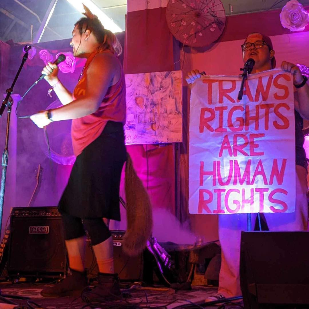 A photo from DreamStates' performance at IngenuityFest 2023; Madeline sings and walks along the stage holding her mic, tail swinging behind her, as Jo stands singing next to her, holding a banner that reads "Trans Rights are Human Rights".