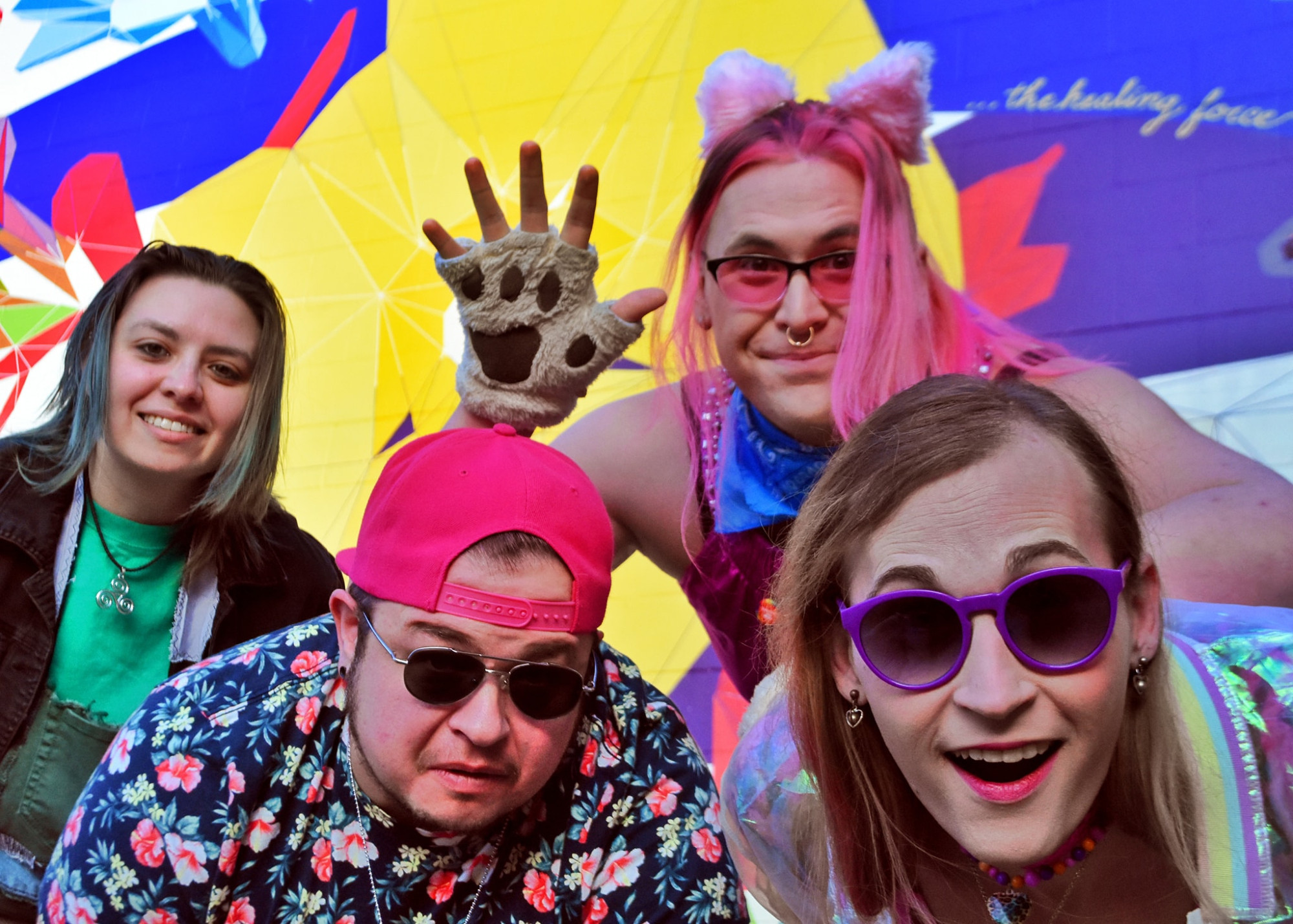 A photo of four transgender musicians standing in front of a mural and looking at the camera. They are dressed in colorful outfits, and a band member wearing fuzzy ears and pawprint gloves is waving.