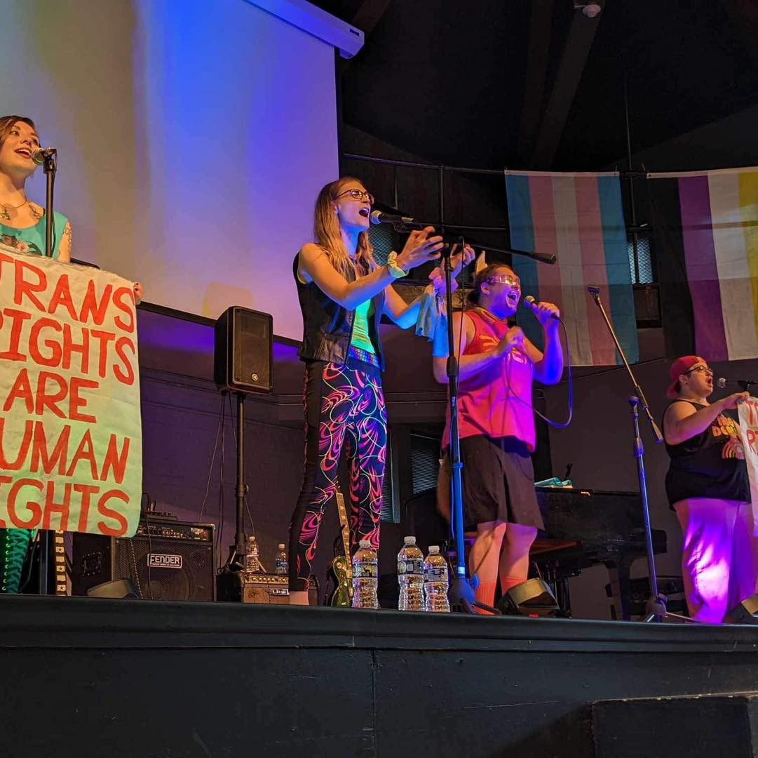 DreamStates onstage at the 14th annual TransOhio Trans and Ally Symposium; Natalie and Madeline are singing together and reaching out to the crowd with their hands, next to them on either side are bandmates Billy and Jo, who are holding up banners that read "Trans Rights are Human Rights".