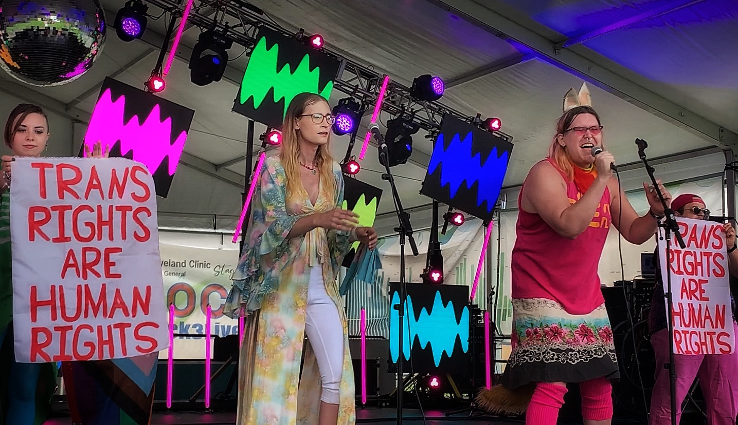 DreamStates on the main stage at Akron Pride 2023; Madeline passionately sings into a mic next to co-lead Natalie, the two of them flanked by bandmates Billy and Jo, who are holding up banners that read "Trans Rights are Human Rights".