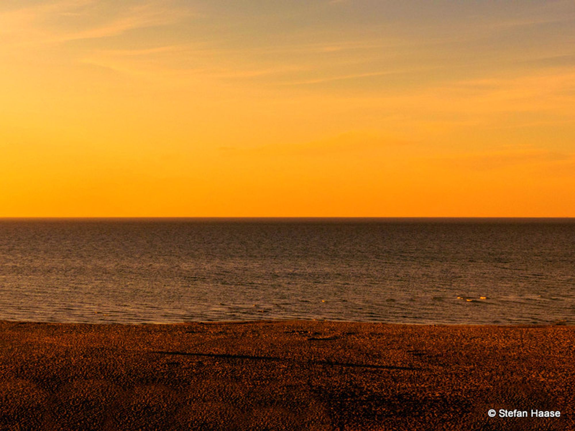 Ein Farbfoto im Querformat. Man sieht einen Strand und der Blick geht hinaus auf das Meer. Das Foto teilt sich in 3 Schichten. Oben die untergehende Sonne, in der Mitte die ruhige See und unten der Strand./ A color photo in landscape format. You can see a beach and the view is out to sea. The photo is divided into 3 layers. The setting sun at the top, the calm sea in the middle and the beach at the bottom.