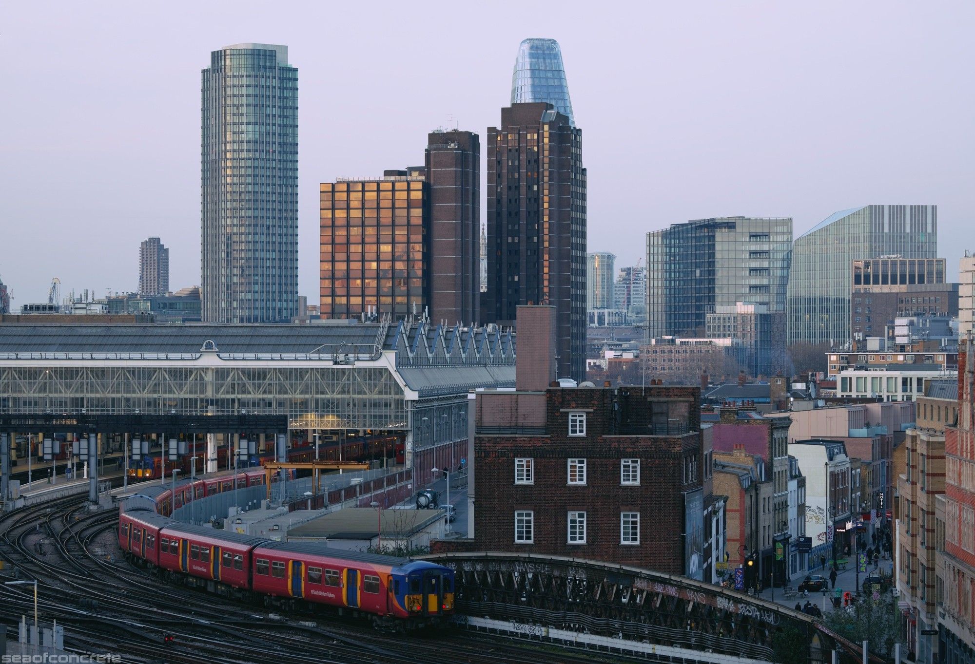 The photo shows the platforms of Waterloo station and the train tracks leading south, with the tower blocks and skyscrapers visible behind it. The photo was taken from the top balcony of Canterbury House, London, SE1.