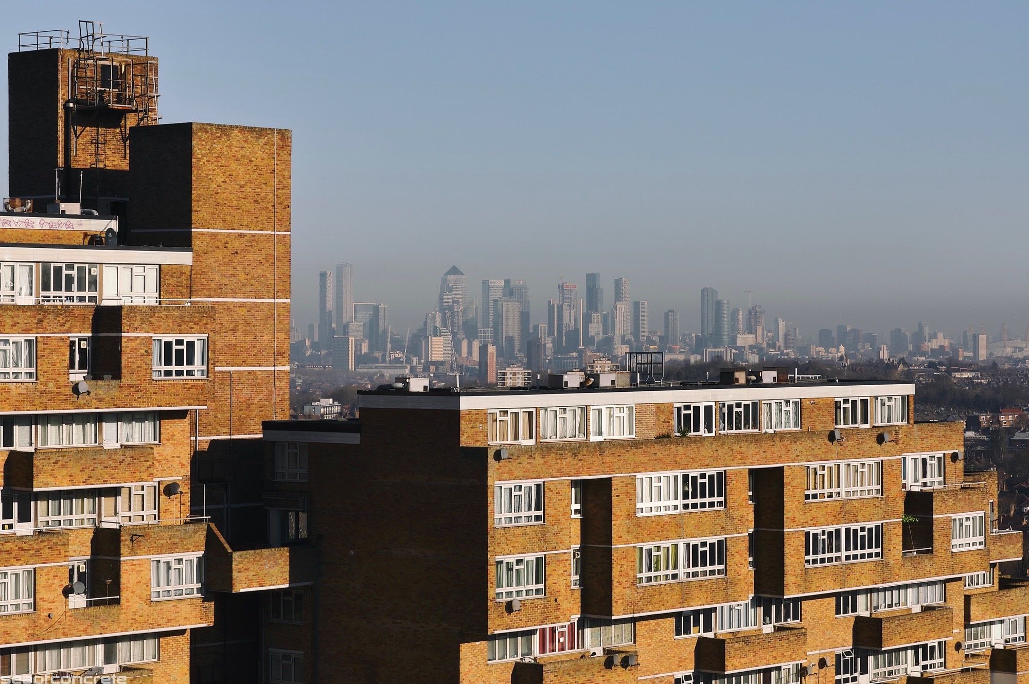 The north block of the Dawson’s Heights estate (called ‘Ladlands’) is illuminated by the early morning sun, with the skyscrapers of Canary Wharf behind it in the distant background. The skies are clear but Canary Wharf is covered by a low-hanging layer of smog.