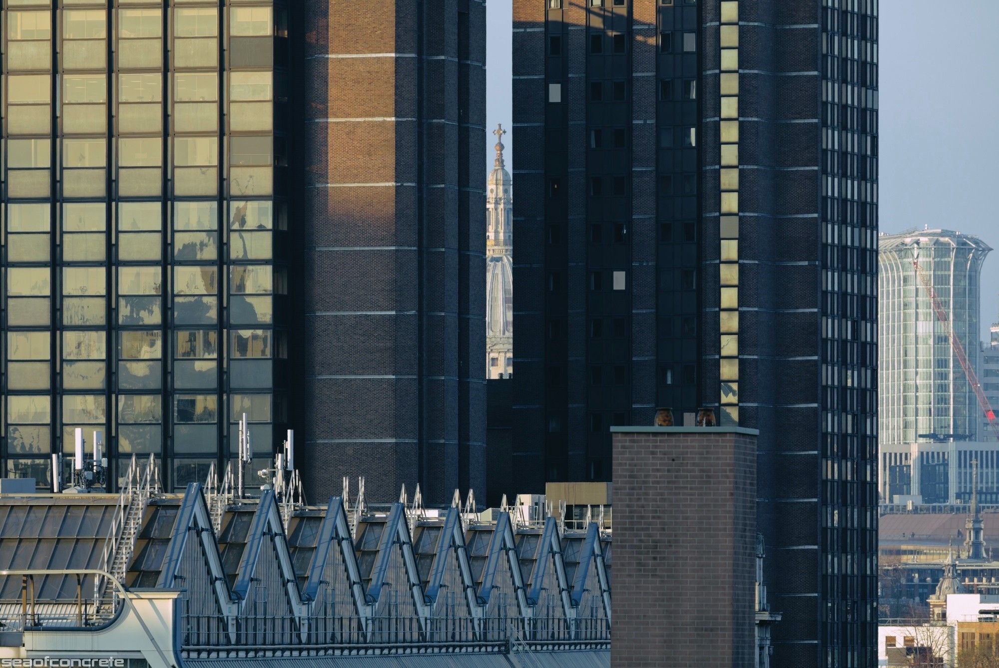 A thin sliver of St. Paul's cathedral, centred on the middle of the dome and with the cross fully visible, can be seen through a gap between two adjacent tower blocks next to Waterloo Station. The jagged roof of the station is also visible in the bottom third of the photo. The photo was taken from Canterbury House, SE1, at sunset in April 2023.