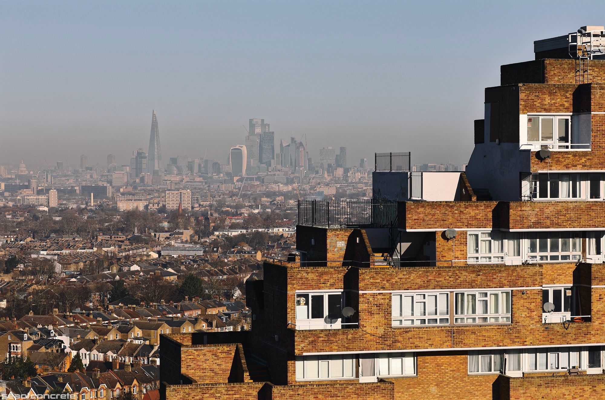 The north block of the Dawson’s Heights estate (called ‘Ladlands’) is illuminated by the early morning sun, with the skyscrapers of the City of London and the Shard behind it in the distant background. The skies are clear but the City of London is covered by a low-hanging layer of smog.