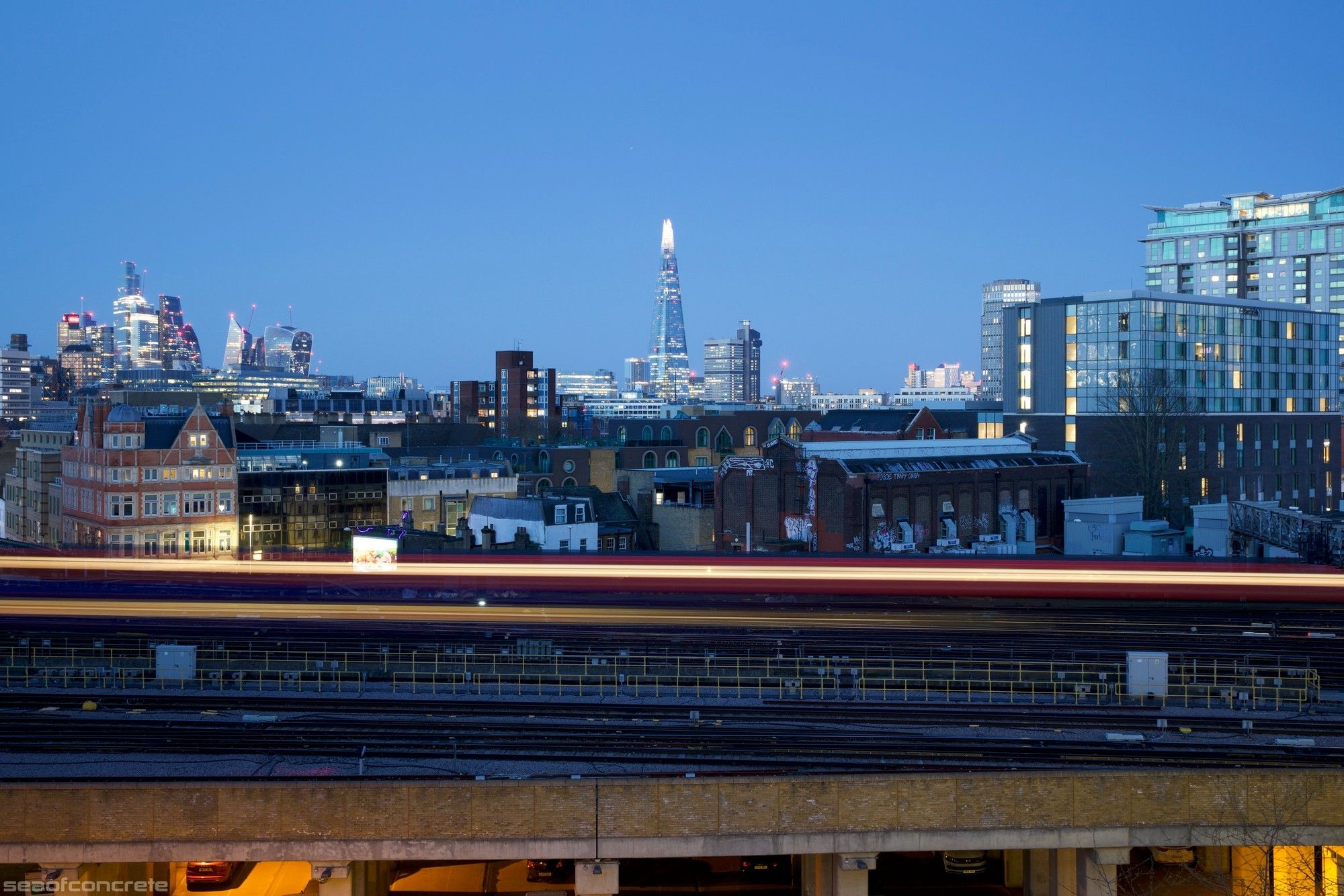 The photo shows the train tracks leading into Waterloo station with the tower blocks and skyscrapers visible behind it. The photo was taken from the top balcony of Canterbury House, London, SE1.