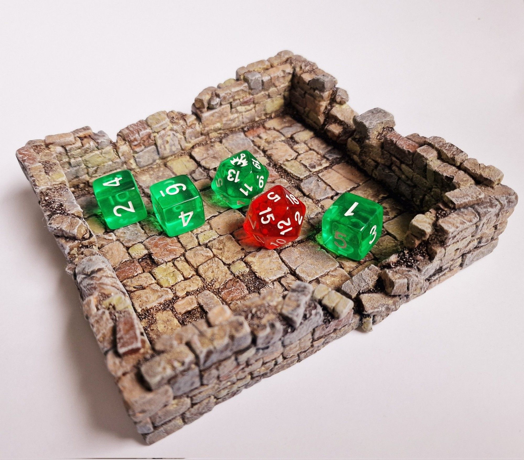 Model of a ruined stone house foundation that's also a dice tray. There's a set of green and red Dragonbane dice showing a critical success.