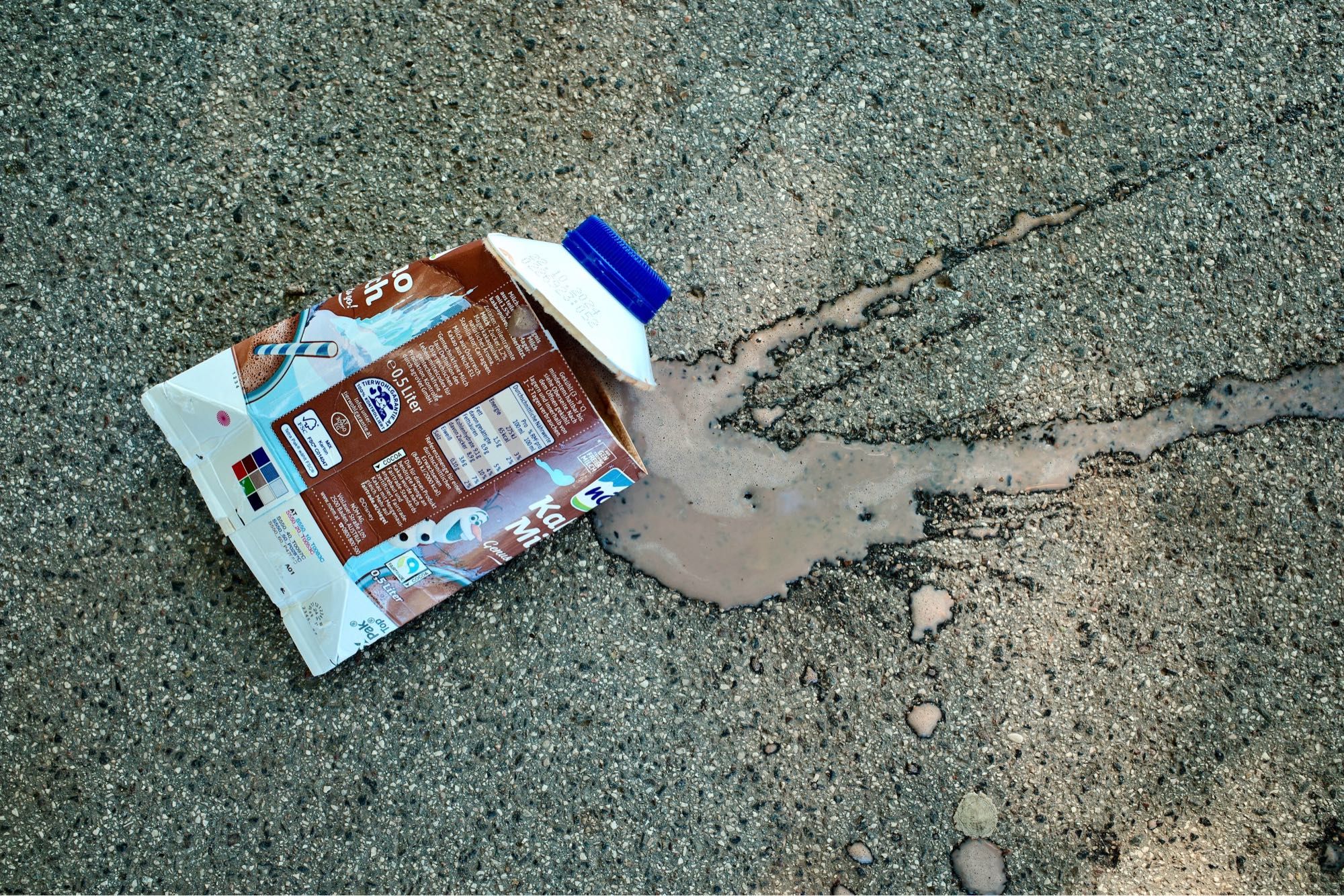 The image shows a spilled carton of chocolate milk lying on an asphalt surface. The carton is partially crushed, with the open top facing sideways, allowing the chocolate milk to flow out and form a small puddle on the ground. The scene captures a moment of accidental waste on a rough street texture.