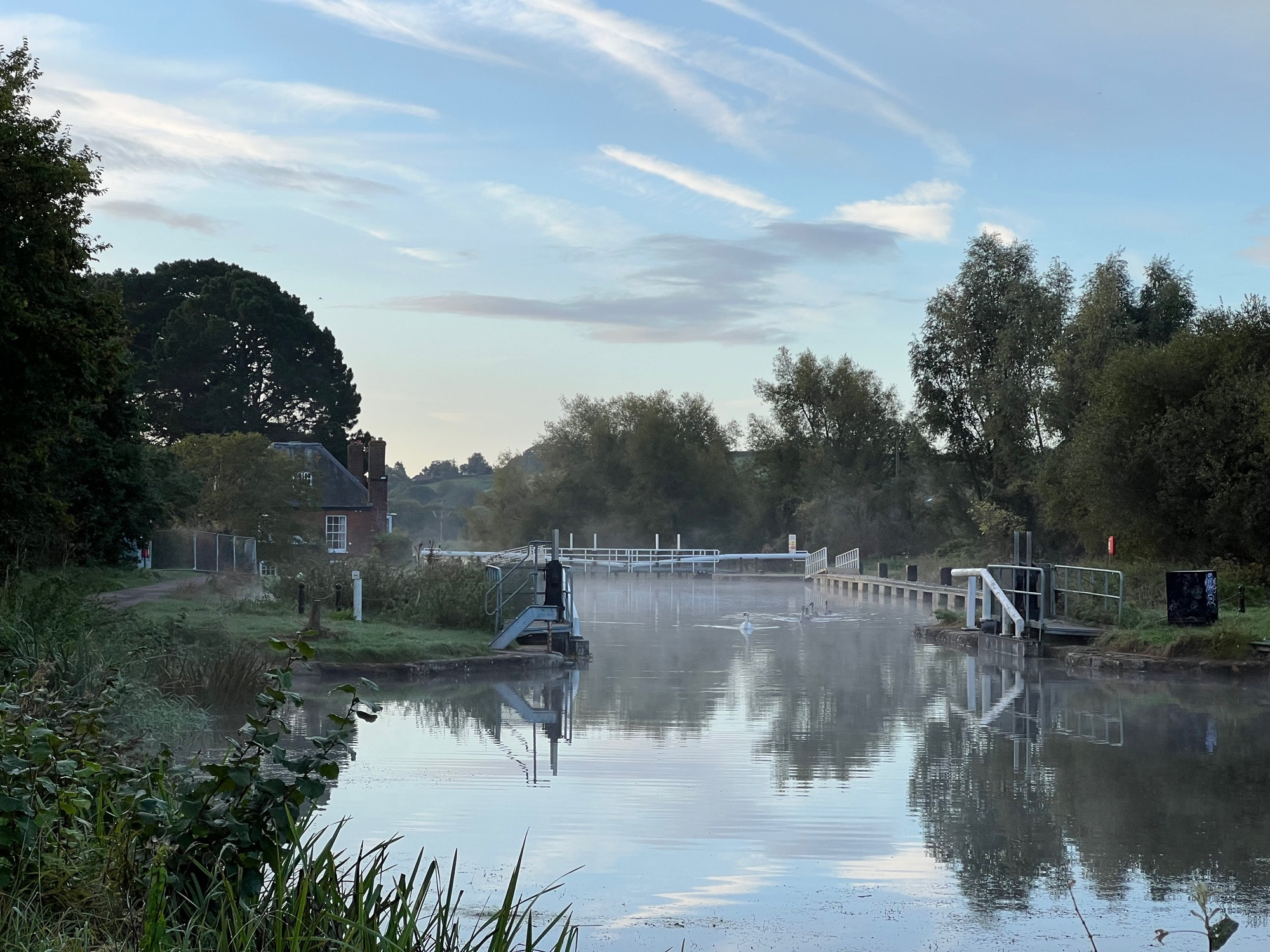 As described in the text, there is a low mist rising over the water, and a family of swans swims towards us in the middle distance. Behind them you can see the lock gates and the Double Locks inn against the tall trees to the left.