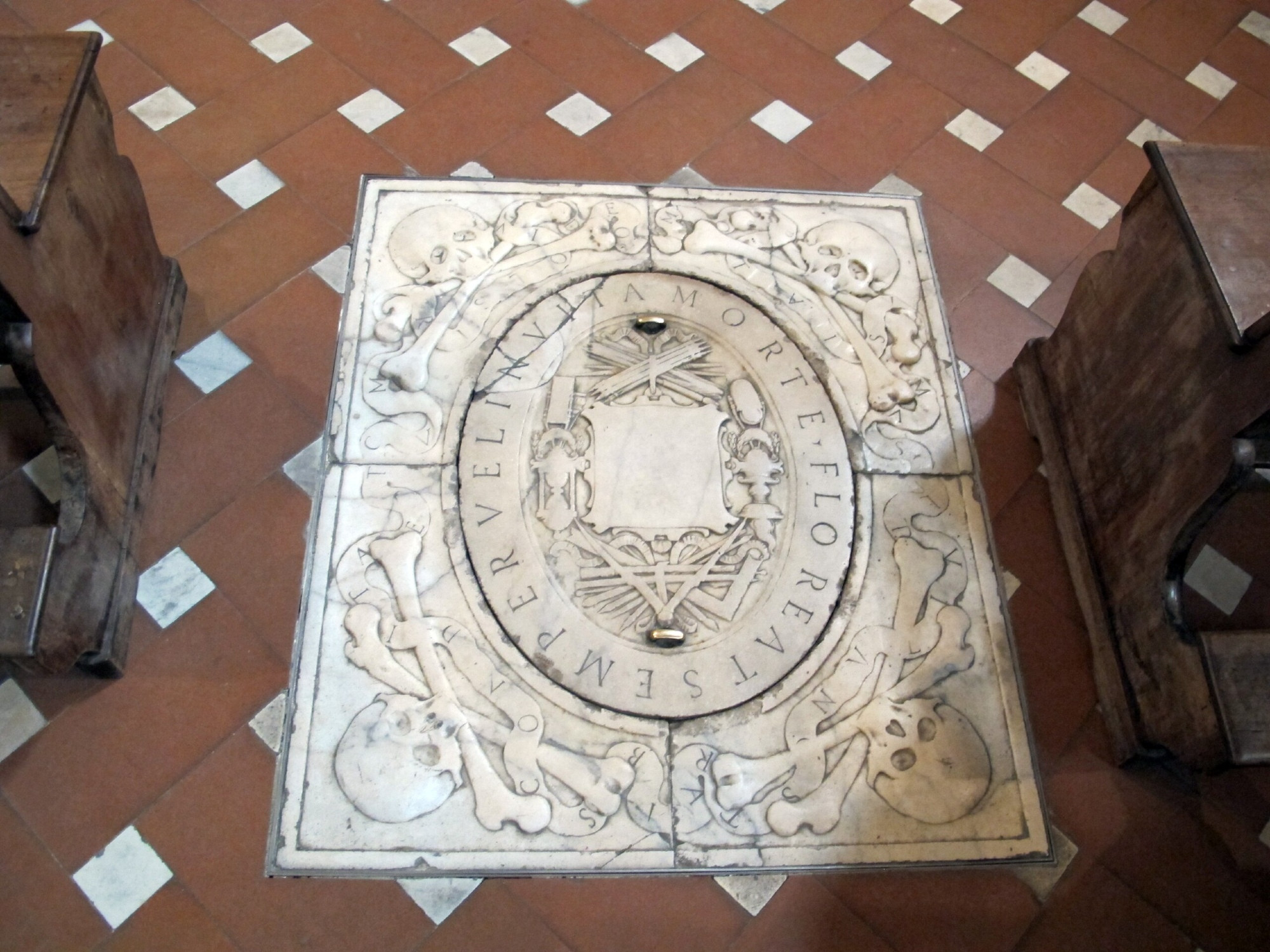 A rectangular marble tomb slab with an oval removable cover at its center. The oval frames a collection of artist's tools like pencils, paint brushes, straight edges, squares and an inscription that reads: FLOREAT SEMPER VEL INVITA MORTE.