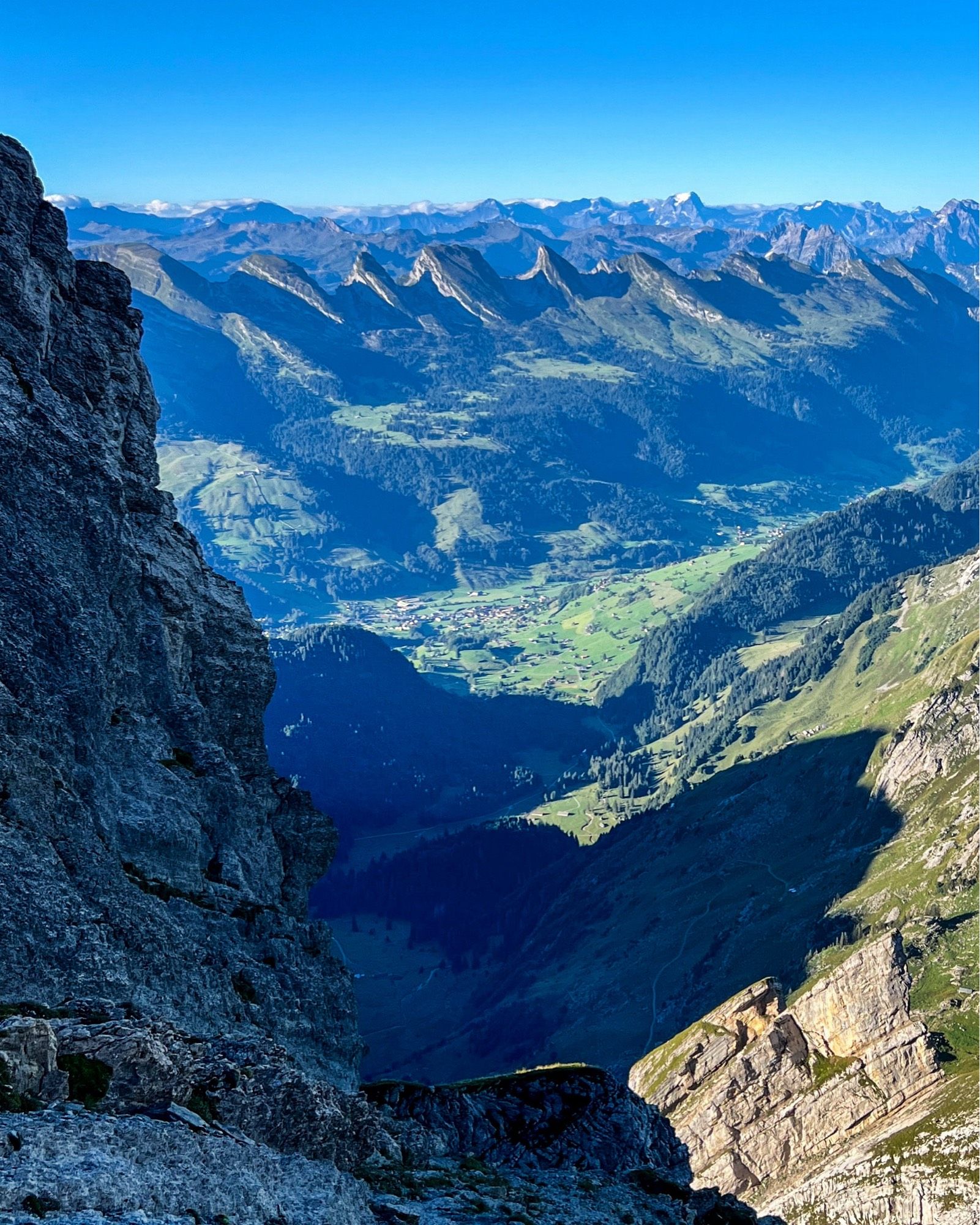 Ein tiefes Tal, das von hohen, felsigen Bergen umgeben ist. Die saftig grünen Wiesen im Talboden bieten einen starken Kontrast zu den grauen Felsen und den dunklen Schatten der Bergwände. Im Hintergrund ist eine Bergkette mit mehreren Gipfeln zu sehen, die sich bis zum Horizont erstrecken.