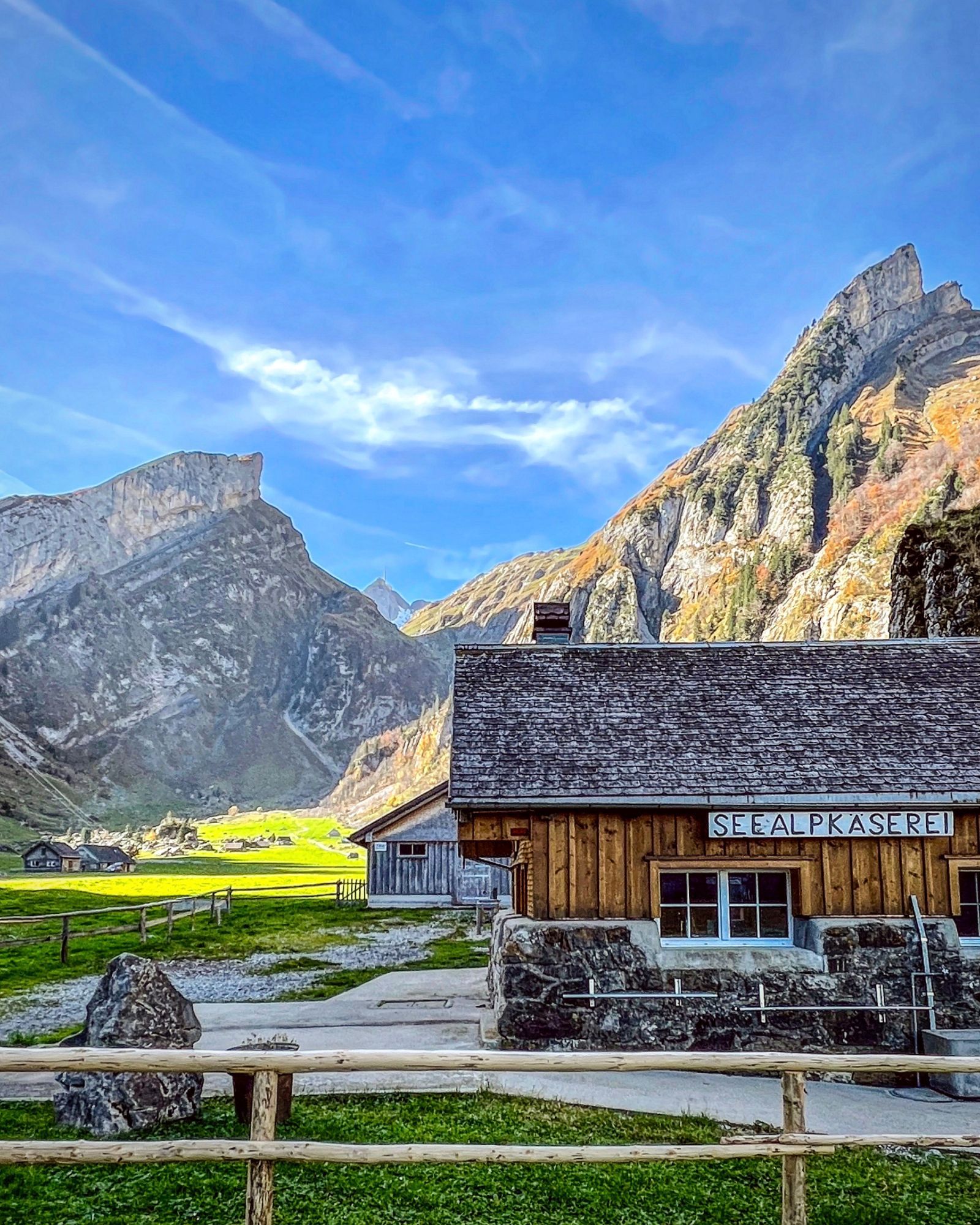 Das Bild zeigt die Seealp in herbstlicher Stimmung im Alpstein. Im Vordergrund steht eine rustikale Holzhütte mit dem Schild "Seealpkäserei". Die umliegenden Wiesen sind noch grün, während die Berghänge in warmen Gelb- und Brauntönen leuchten. Im Hintergrund erheben sich steile, felsige Berge unter einem strahlend blauen Himmel mit ein paar dünnen Wolken.