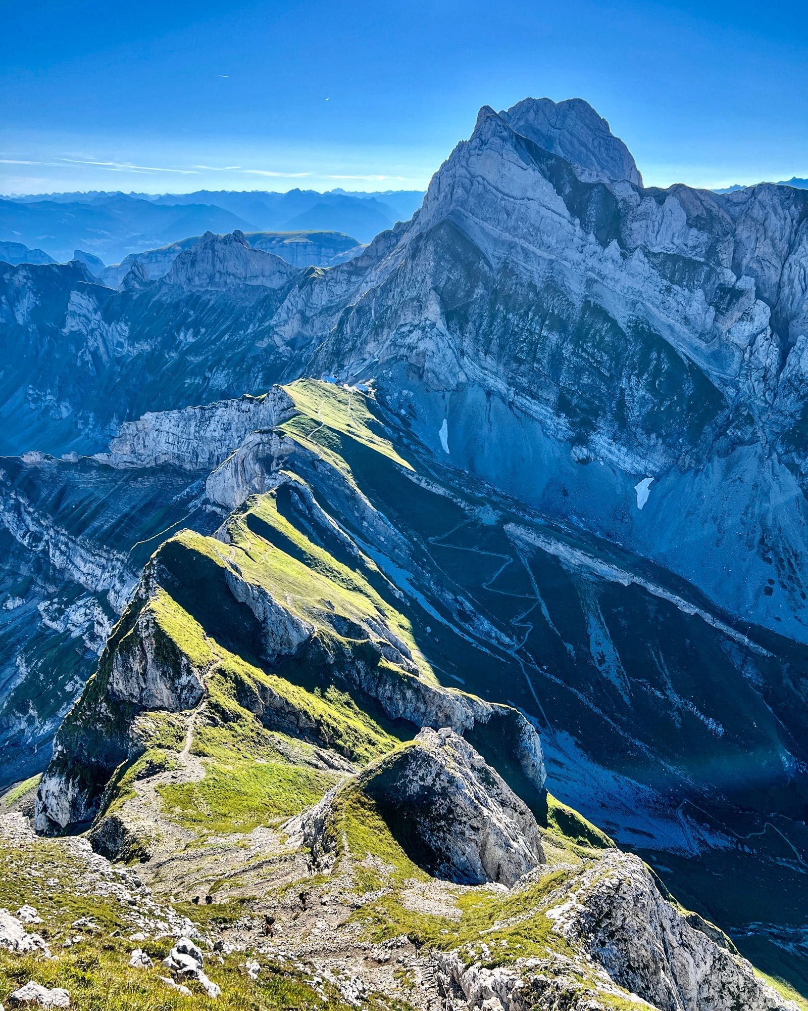 Ein atemberaubender Blick auf eine zerklüftete Bergkette, die von saftig grünen Wiesen auf den Bergkämmen durchzogen wird. Im Hintergrund erhebt sich ein mächtiger, felsiger Gipfel unter klarem, blauem Himmel. Die verschiedenen Schattierungen der Berge und Täler verleihen dem Bild eine beeindruckende Tiefe.