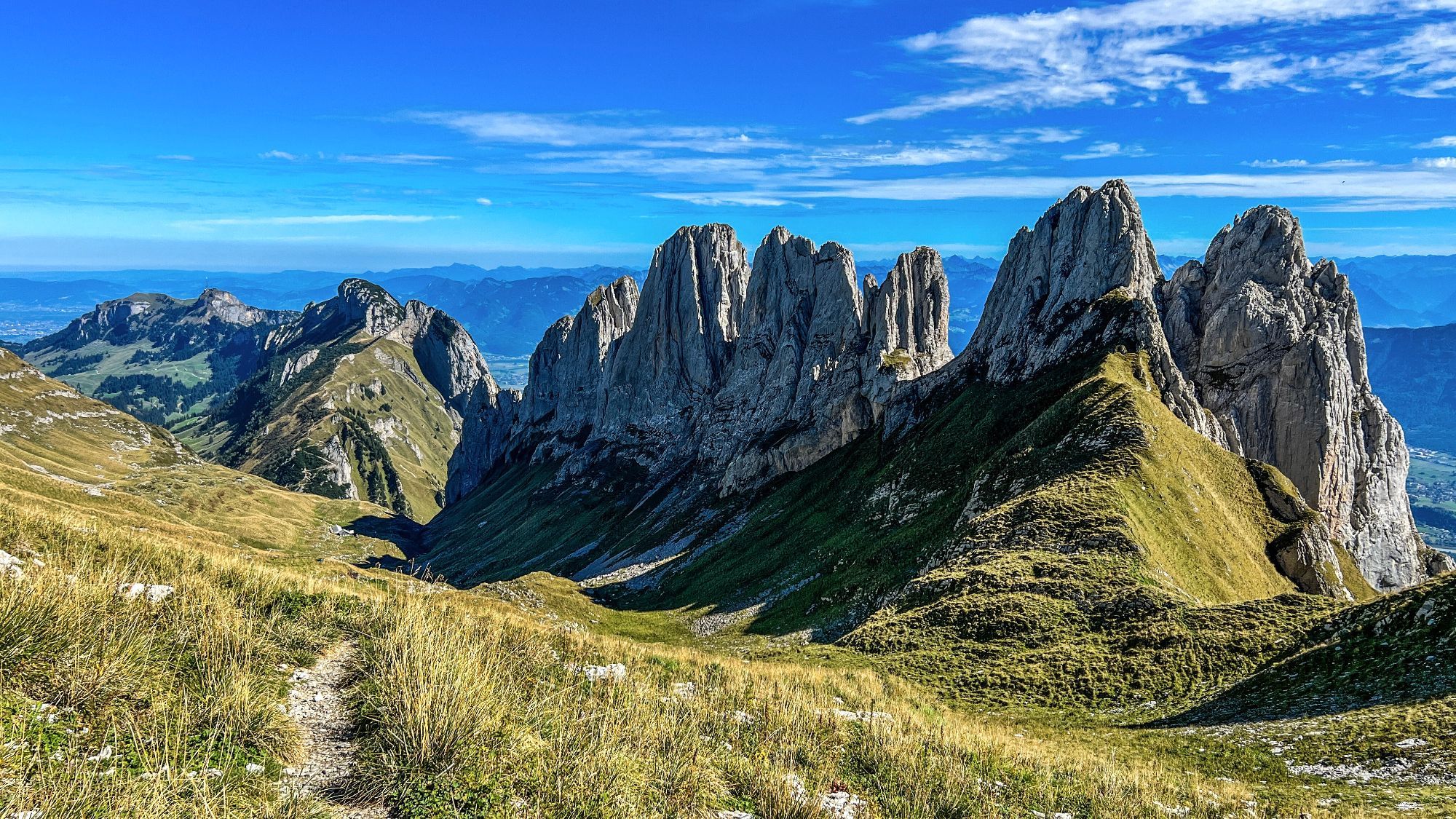 Das Foto zeigt eine eindrucksvolle Berglandschaft mit einer steilen Felskette, die sich in die Höhe erstreckt. Im Vordergrund verläuft ein schmaler, steiniger Wanderweg durch eine grasbewachsene Alpwiese. Im Hintergrund erstrecken sich die markanten Gipfel der Kreuzberge im Alpstein-Gebirge, die in steilen, schroffen Felsen aufragen. Der Himmel ist strahlend blau mit einigen dünnen Wolken. Rechts von der Felswand sind grüne Hänge und Täler sichtbar, die in die Ferne reichen. Die Landschaft wirkt ruhig und majestätisch, mit klaren Sichtlinien und einem weiten Blick auf die umliegenden Berge.