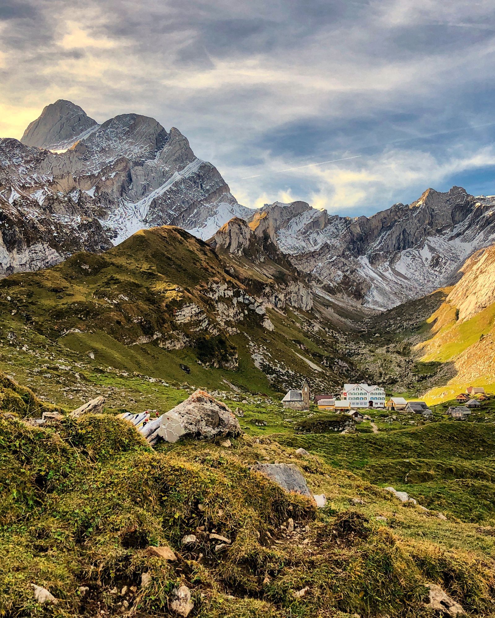 Auf dem Bild sieht man eine malerische alpine Landschaft bei Sonnenuntergang. Im Vordergrund erstrecken sich grüne, mit Gras und Felsen bedeckte Hügel. Im Hintergrund erheben sich imposante, schneebedeckte Berggipfel, darunter der prominente Gipfel des Altmanns (links). Weiter rechts ragen die schroffen, steinigen Felswände des Alpsteingebirges in die Höhe. Am unteren Rand des Bildes befindet sich die Meglisalp, eine kleine Siedlung mit traditionellen Berghäusern, die in der malerischen Berglandschaft eingebettet ist. Die Stimmung ist ruhig, und der Himmel ist von sanften Wolken bedeckt, durch die die tief stehende Sonne goldenes Licht wirft.