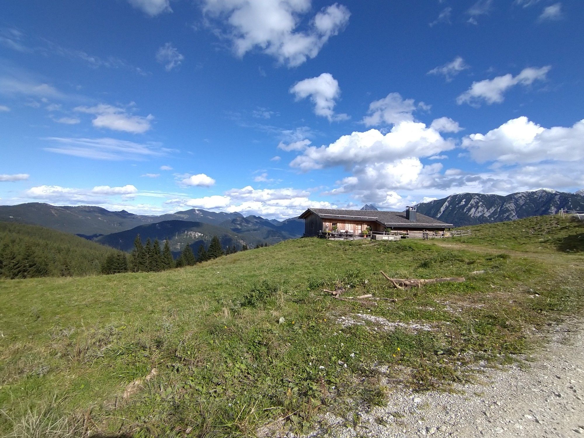 Blick von einer Alm. Man sieht Wiesen und eine Hütte im Vordergrund, dahinter teils waldige Täler und hohe Berge am Horizont.