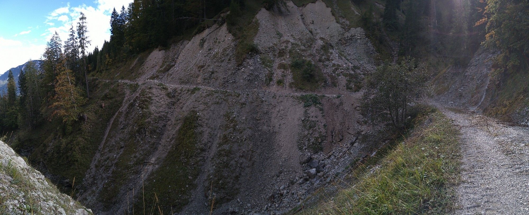 Panoramafoto aus den Alpen. Man sieht, wie der Weg etwa einen 180-Grad-Bogen beschreibt, sozusagen ein U mit der Öffnung nach links. Links des Wegs geht es steil bergab, rechts ziemlich steil bergauf, teils ist der Hang abgestürzt. Ganz rechts fließt Wasser nach unten und über den Weg. Am linken Bildrand reicht der Blick vorbei an Nadelbäumen bis zum bergigen Horizont unter einem blauen Himmel mit vereinzelten Wölkchen.