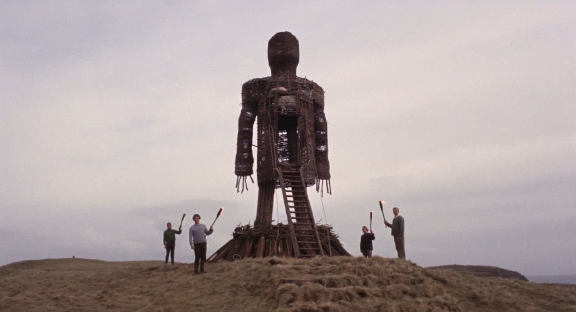 a giant wicker man effigy in an open field towers over the men standing around it who are each holding aloft their lit torches