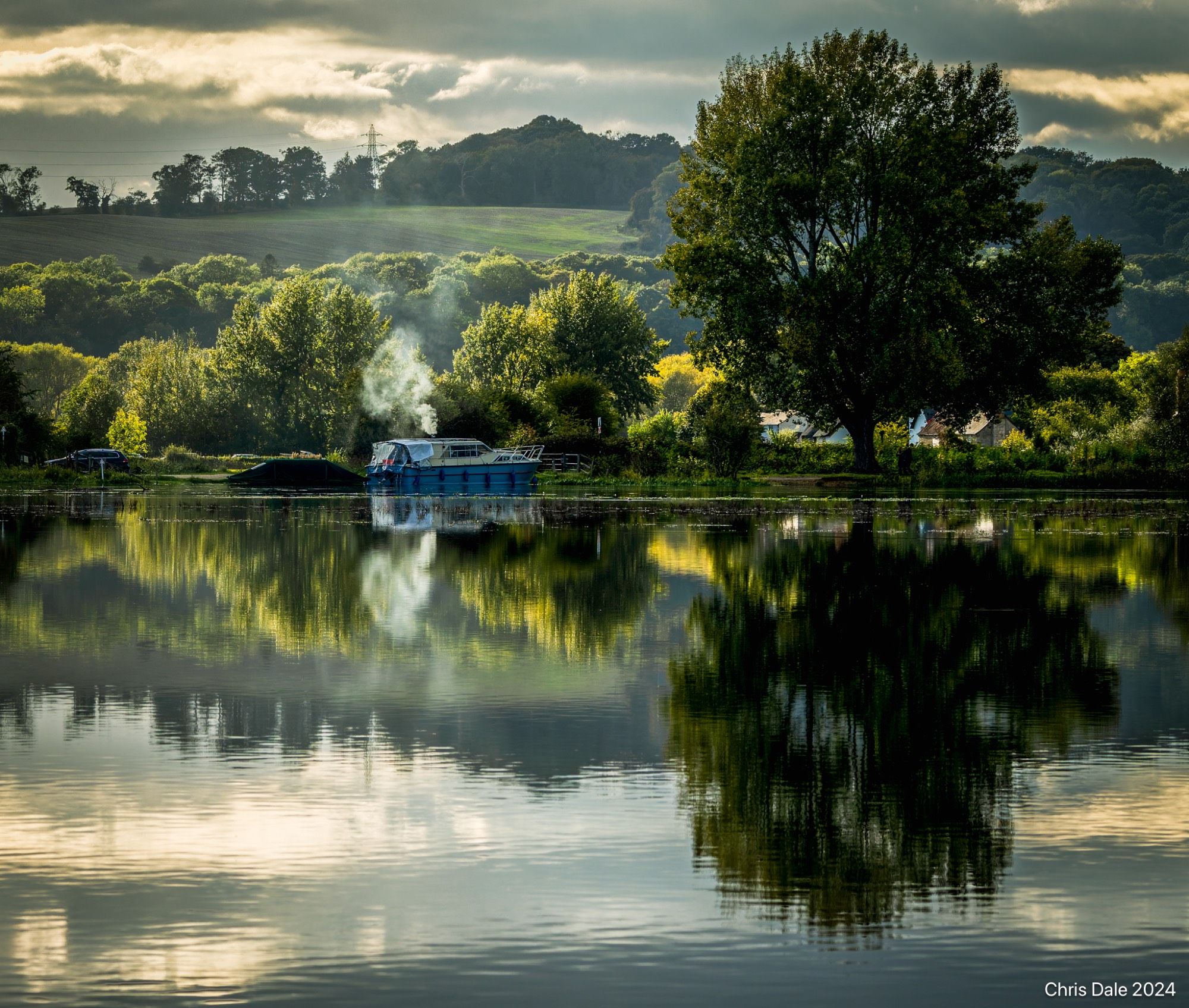 View across still water to trees and a hill beyond. A column of smoke rises behind a boat on the river