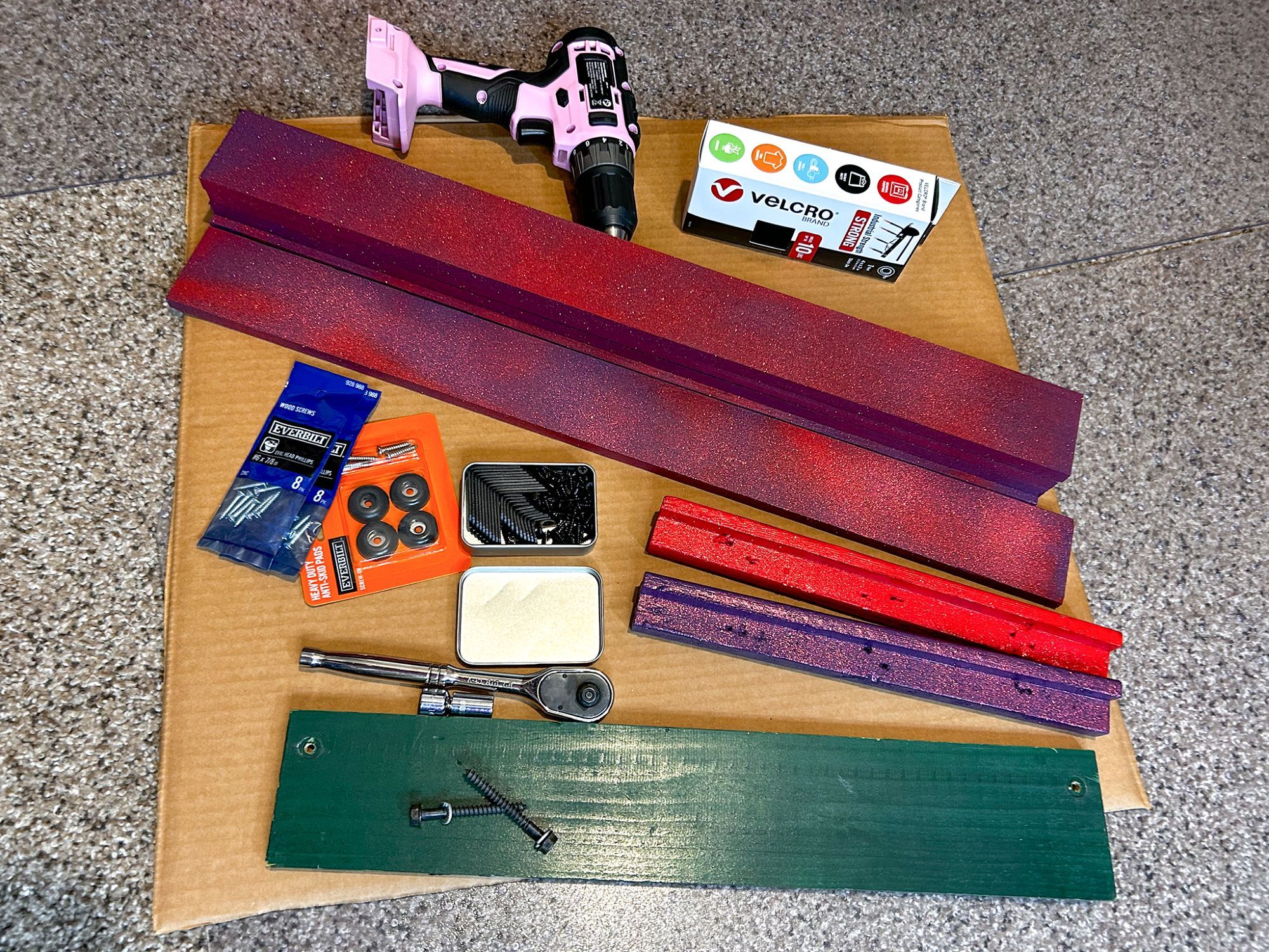 All boards colored & dried, then a drill, box of velcro, bag of screws, screw-on rubber feet, L-shaped brackets, a ratchet with a socket, and then two screws laying on the old backboard. They are all sitting on a piece of cardboard