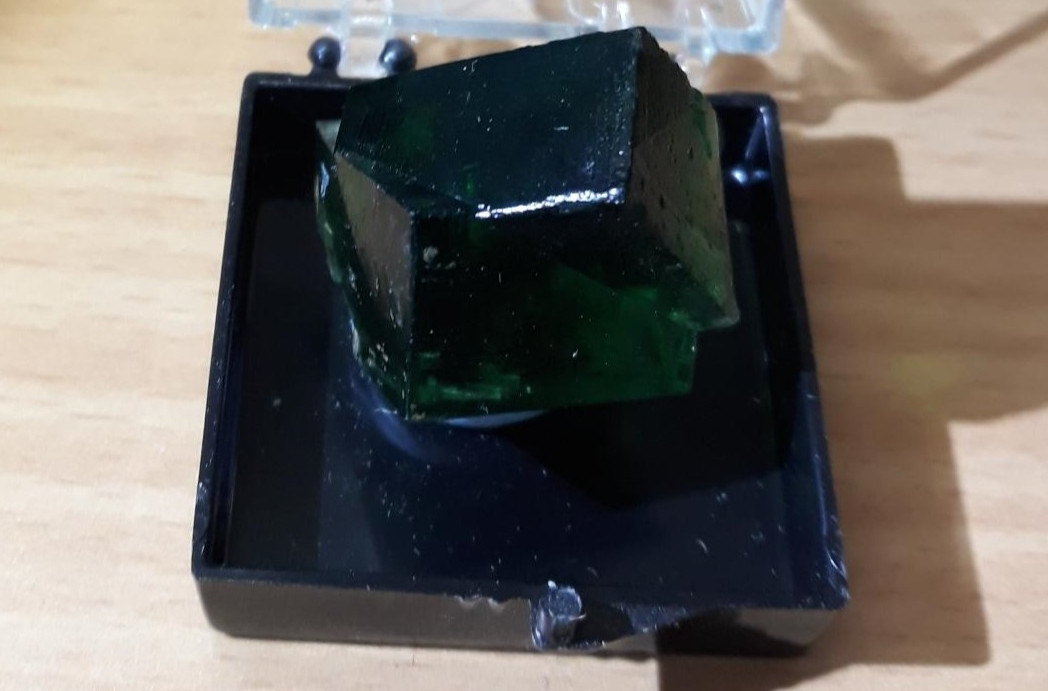 Small, translucent, dark green fluorite in an open plastic box with a hinged lid. The fluorite shows twinning so it looks like two interlocking cubes. Specimen is from Rogerley Mine, County Durham, England. From the Natural History Museum, London's collection.