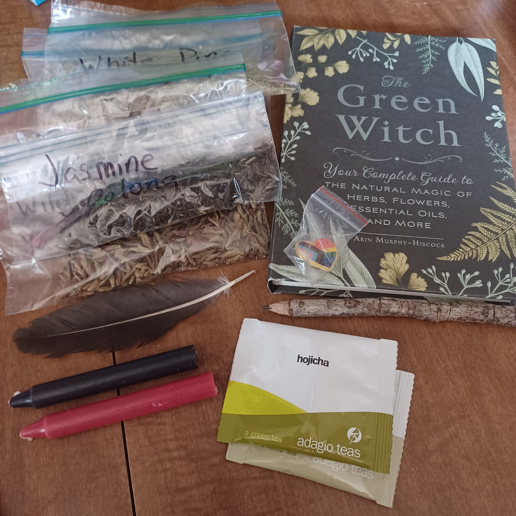 A picture of various items including a red and black candle, a black bird feather, ziploc bags with various herbs and tea blends stacked together with some visible saying jasmine oolong and another saying white pine, then there is a book titled The Green Witch by Arin Murphy-Hiscock.