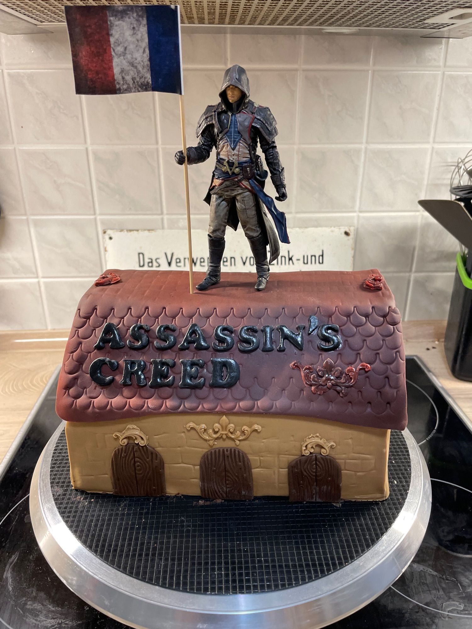 Kuchen in Form eines alten Hauses. Auf dem Dach steht Assasin’s Creed. Oben drauf eine Figur von Arno Dorian.