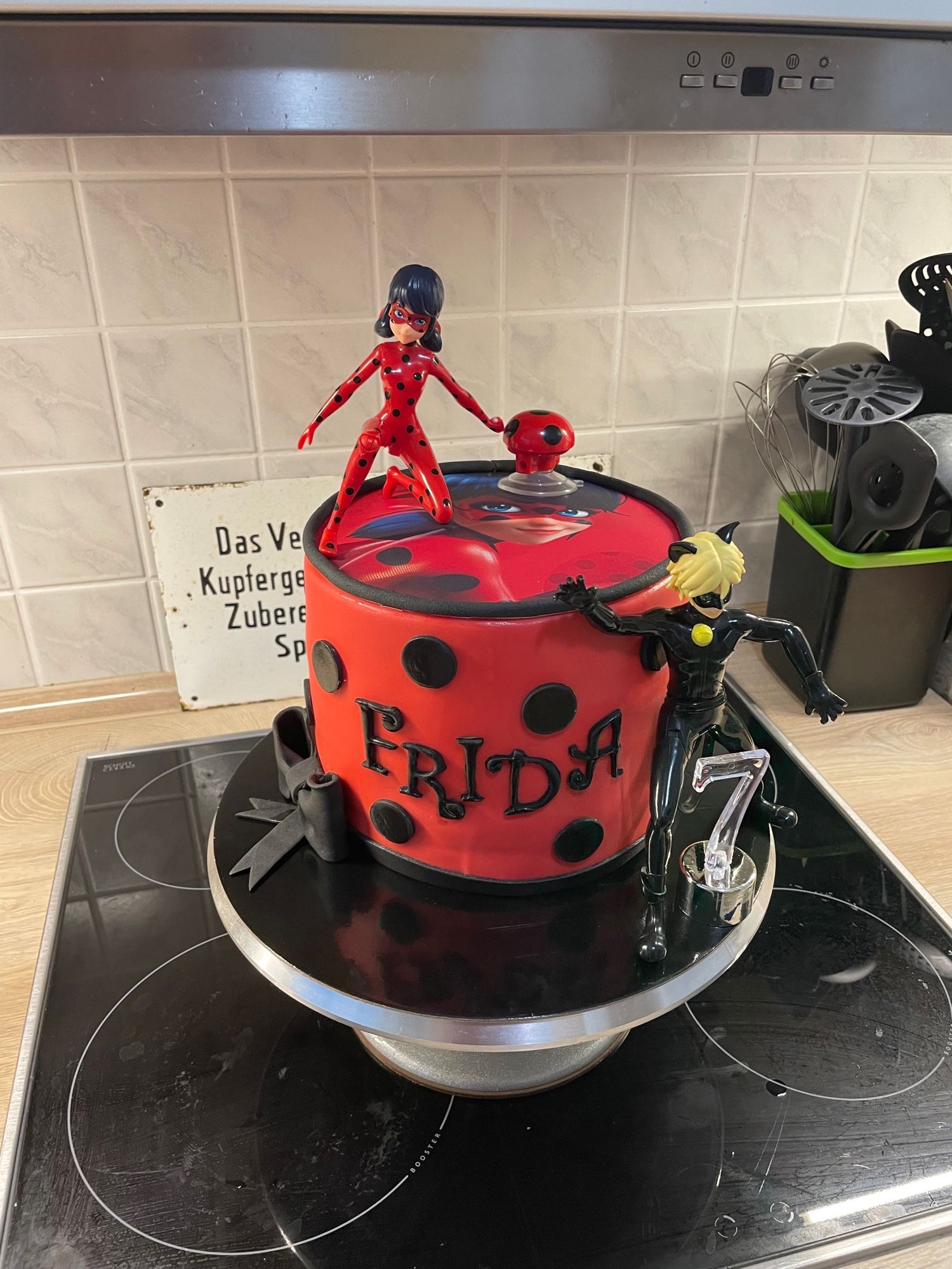 Torte im Lady Bug Design, d.h. rot mit schwarzen Punkten. Oben drauf ein Bild und eine Figur von Lady Bug. An der Seite eine Figur von Cat Noir.
Für nicht eingeweihte: es handelt sich um eine Zeichentrickserie für Kinder. Ein Mädchen im Marienkäferkostüm rettet die Welt … oder so ähnlich … ich weiß es doch auch nicht …