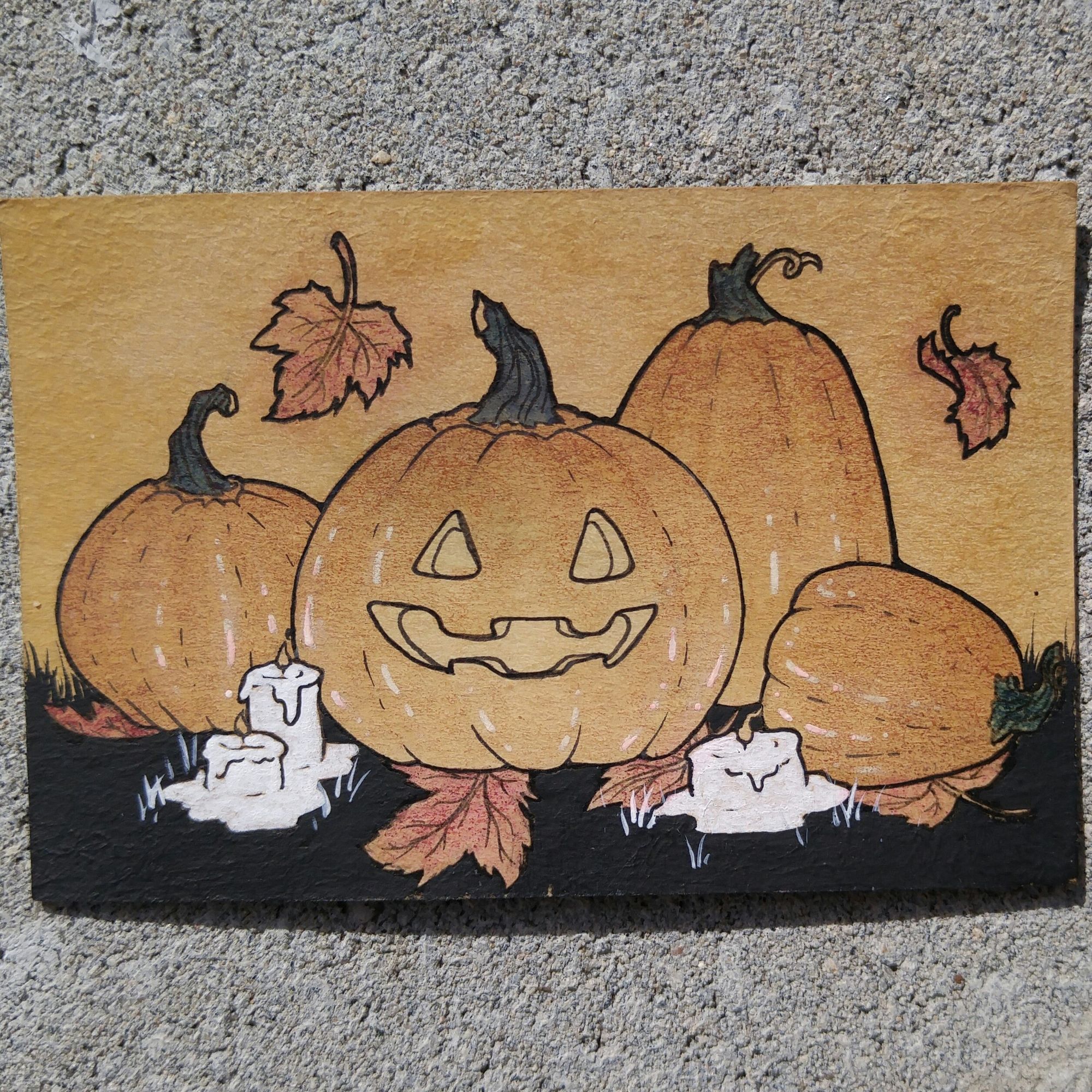 A photo of a trading card sized mixed media art card (ink, acrylic paint, marker, gel pen, on tea-stained cardstock). The art depicts four orange pumpkins of various shapes and sizes. The largest, in the center, has a smiling jack-o'-lantern face. The smallest, rightmost pumpkin is tipped over on its side. There are also six maple leaves scattered around and three lit white wax candles.