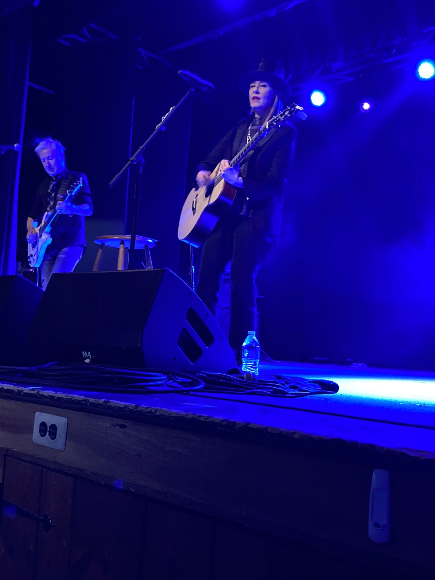 Suzanne Vega playing guitar and singing. She’s wearing a black jacket, a top hat, and jeans.