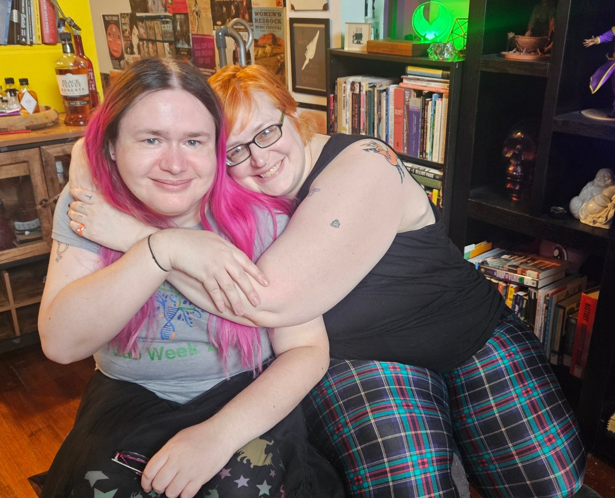 on the right, a white trans woman with long pink hair wearing a gray shirt smiles and is being hugged by a white nonbinary person with short, bright orange hair and black glasses, wearing a black tank top.