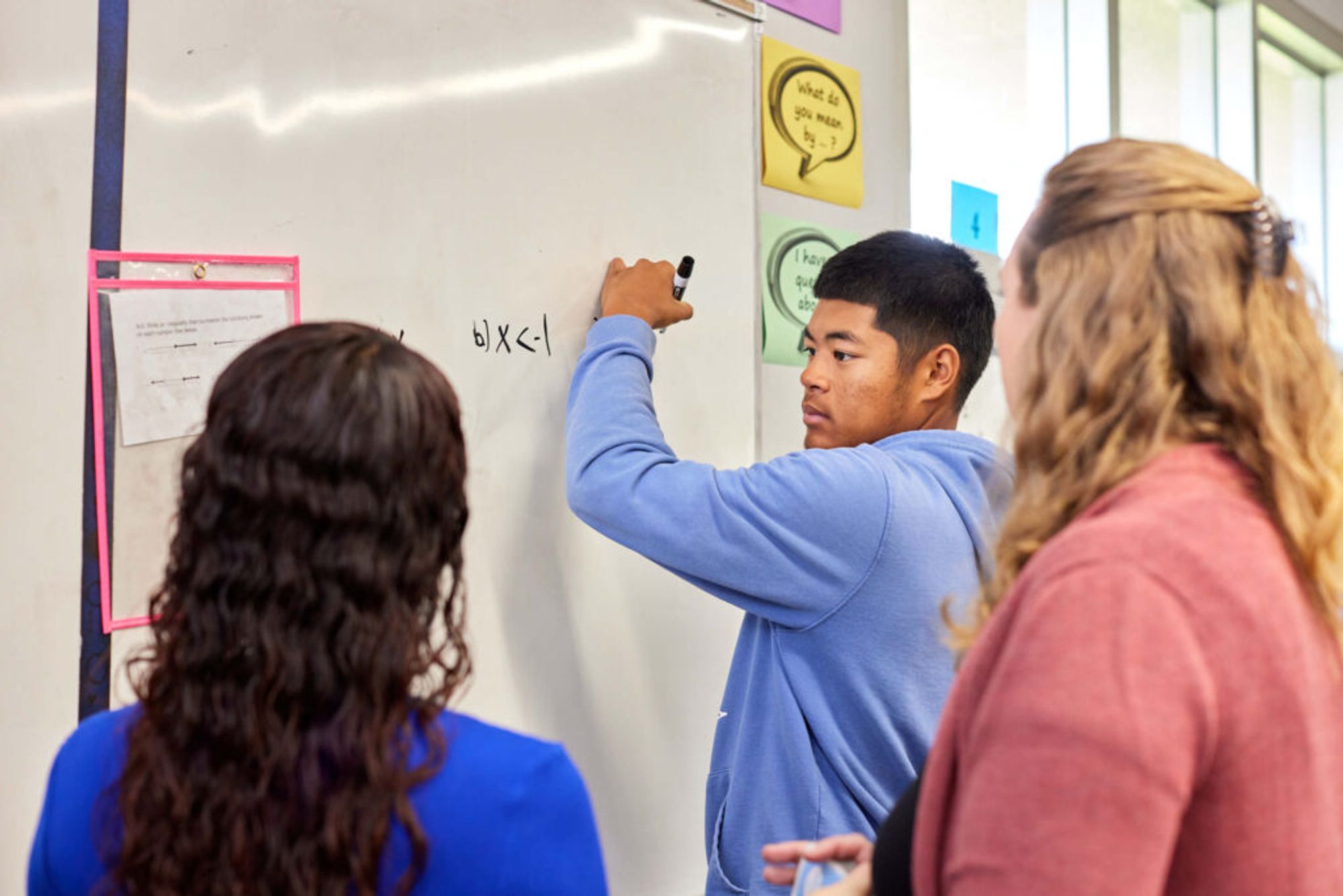 Three people in a classroom looking at a whiteboard that reads “b) x < –1.”