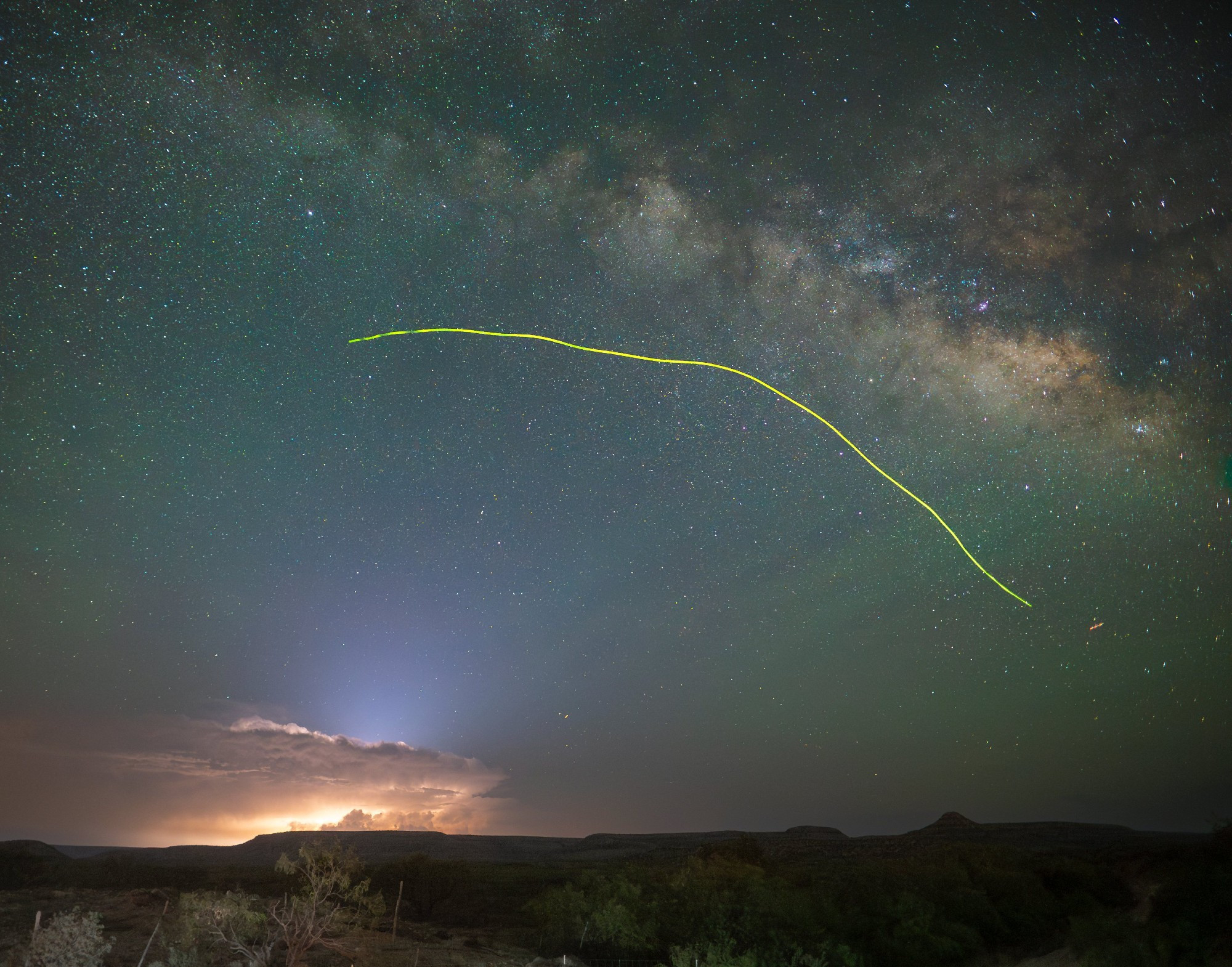 L'image montre un ciel étoilé au-dessus d'un paysage naturel.
A l'horizon, des nuages d'orages sont illuminés de l'intérieur et une faible luminosité blanchâtre en sort au-dessus.
Dans le ciel : la voie lactée et des milliers d'étoiles, et au milieu du ciel, une longue trace fine et jaune, irrégulière.