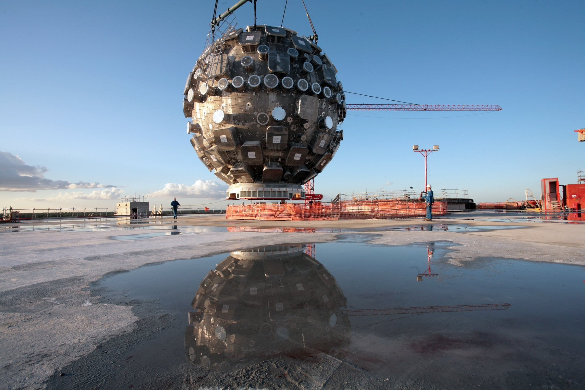 Suspendue au-dessus d'un sol bétonné mouillé de flaques, une structure métallique circulaire est suspendue à des câbles. Elle est constellée de structures carrées et circulaires ressemblant à des hublots ou des trous bouchés.
Au loin, un paysage de travaux avec des structures bétonnées et des grues.