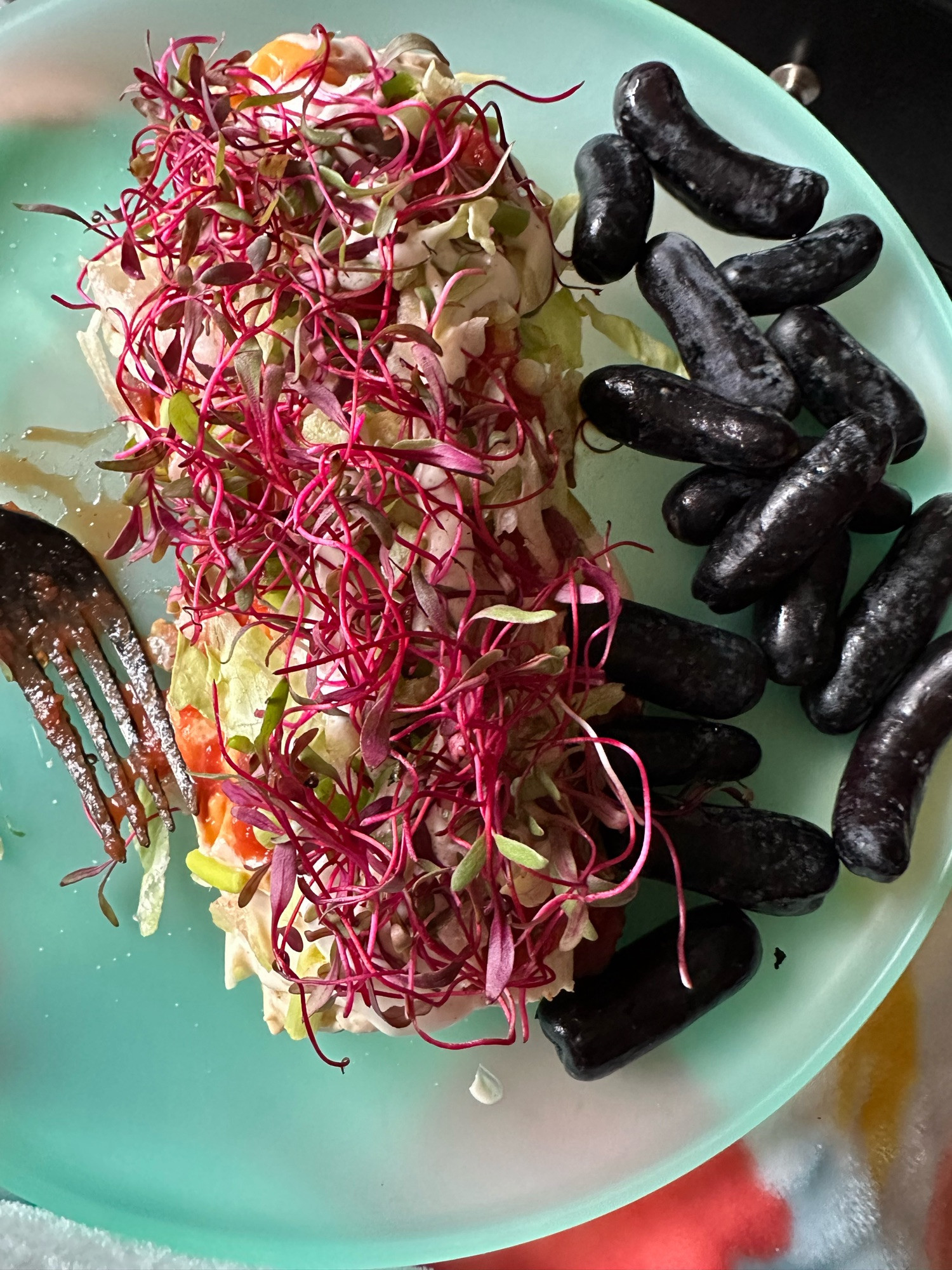 A bean and cheese burrito covered in two cheese sauces: one with queso and one nacho, mild chunky salsas chopped green onions, slivered lettuce, ranch dressing, beet root microgreens, and a side of moon drop grapes which are oblong tubes.