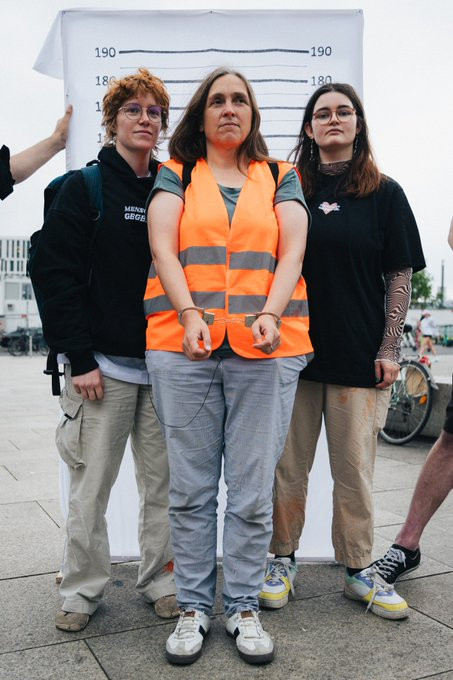 Mensch in oranger Weste und in Handschellen steht vor Mugshot-Wand