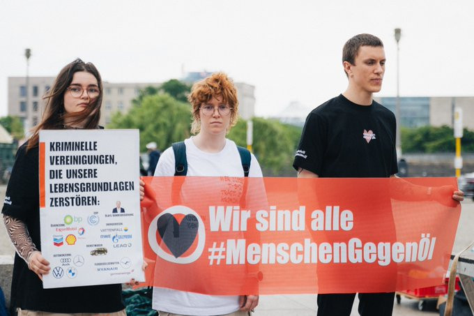 Menschen halten Banner mit der Aufschrift "Wir sind alle #MenschenGegenÖl". Ein Mensch hält ein Plakat mit der Aufschrift "Kriminelle Vereinigungen, die unsere Lebensgrundlagen zerstören:". Darunter sind Logos von fossilen Firmen, u.a. Shell, bp, VW