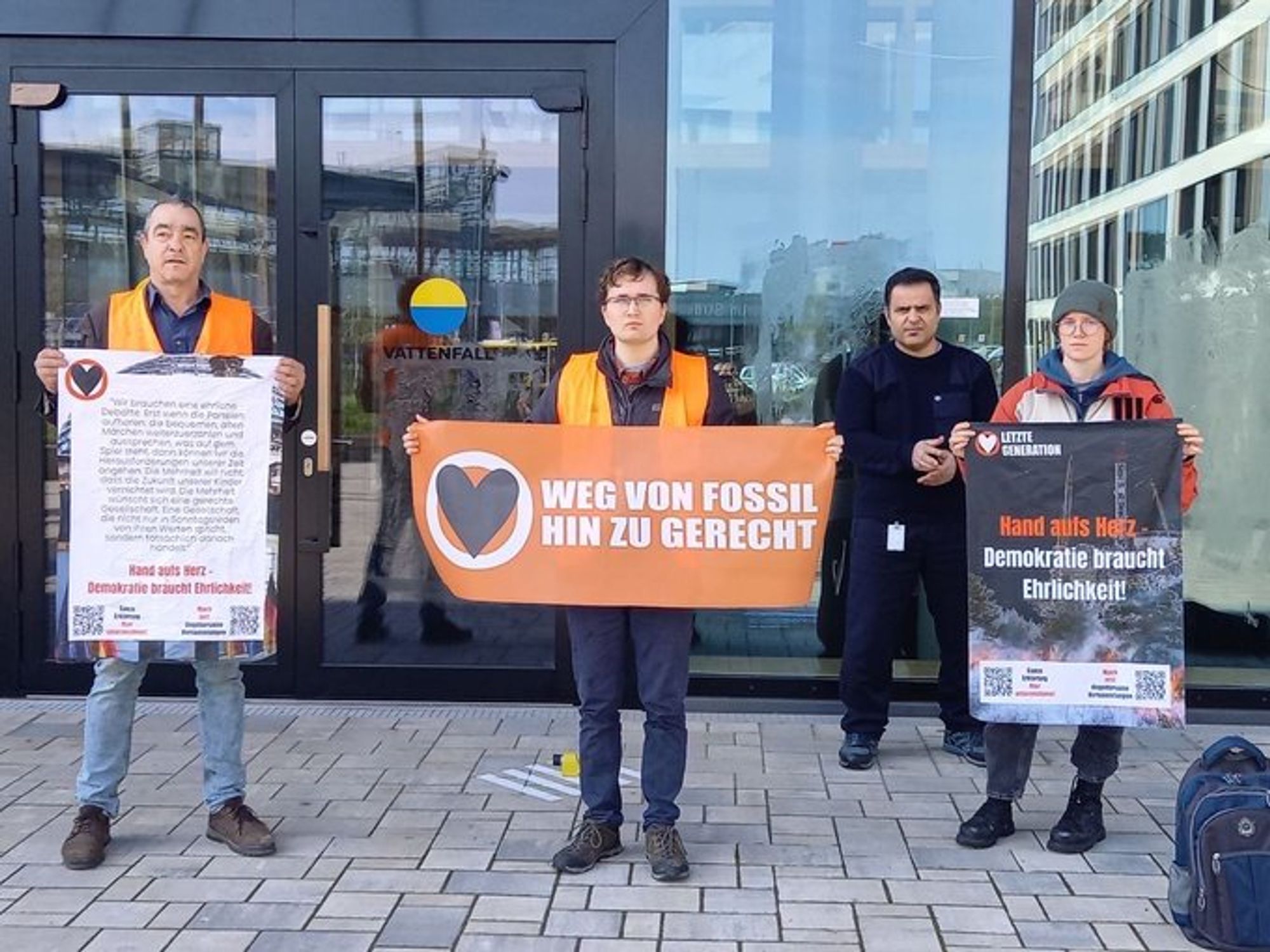 Menschen stehen mit Plakaten vor dem Vattenfall-Gebäude. Auf den Plakaten steht: "Weg von Fossil, Hin zu Gerecht",  "Hand aufs Herz - Demokratie braucht Ehrlichkeit"