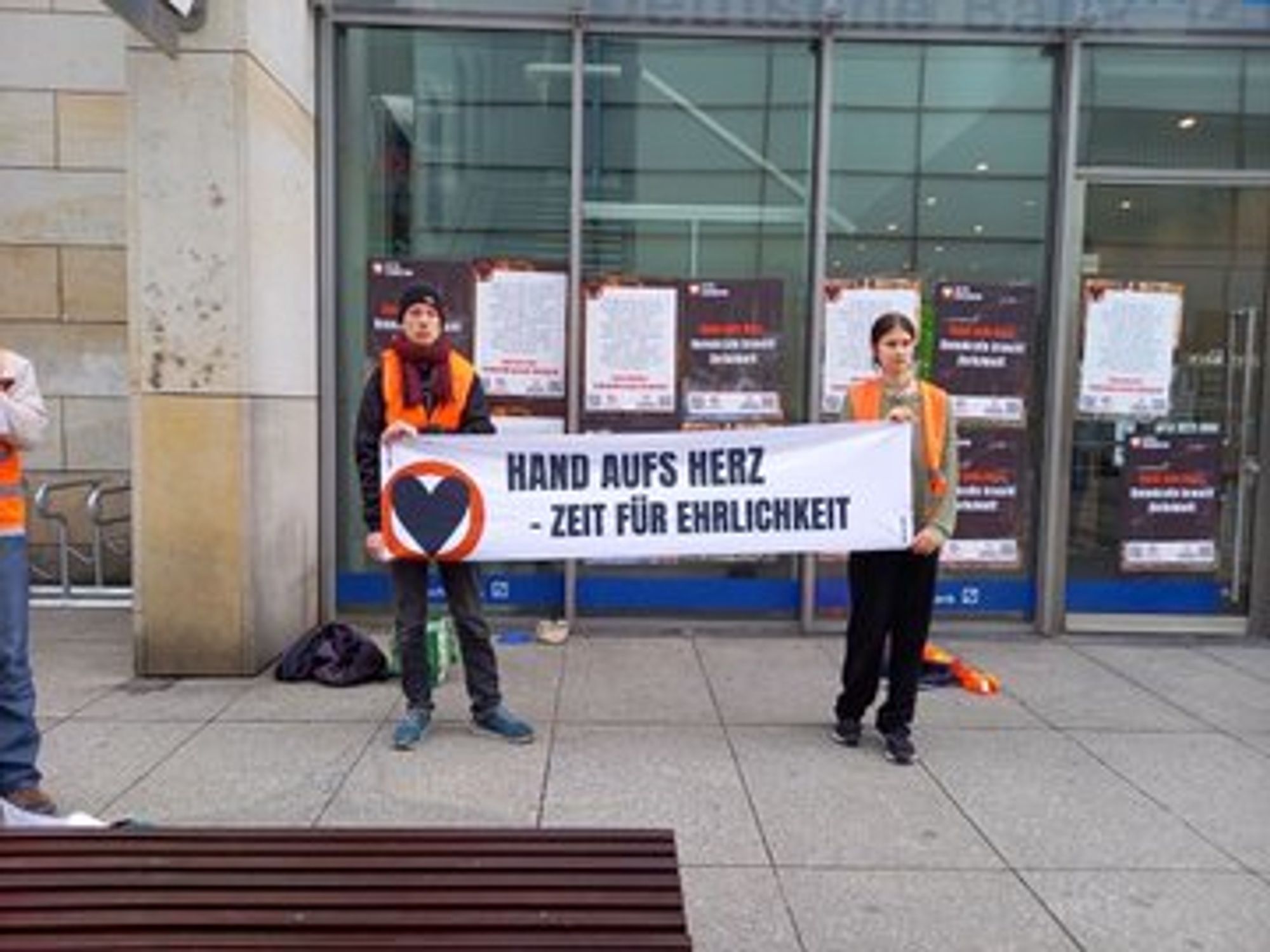 Menschen stehen mit einem Banner mit Aufschrift "Hand auf's Herz - Zeit für Ehrlichkeit" vor einer Filiale der Deutschen Bank. An der Bank kleben Plakate