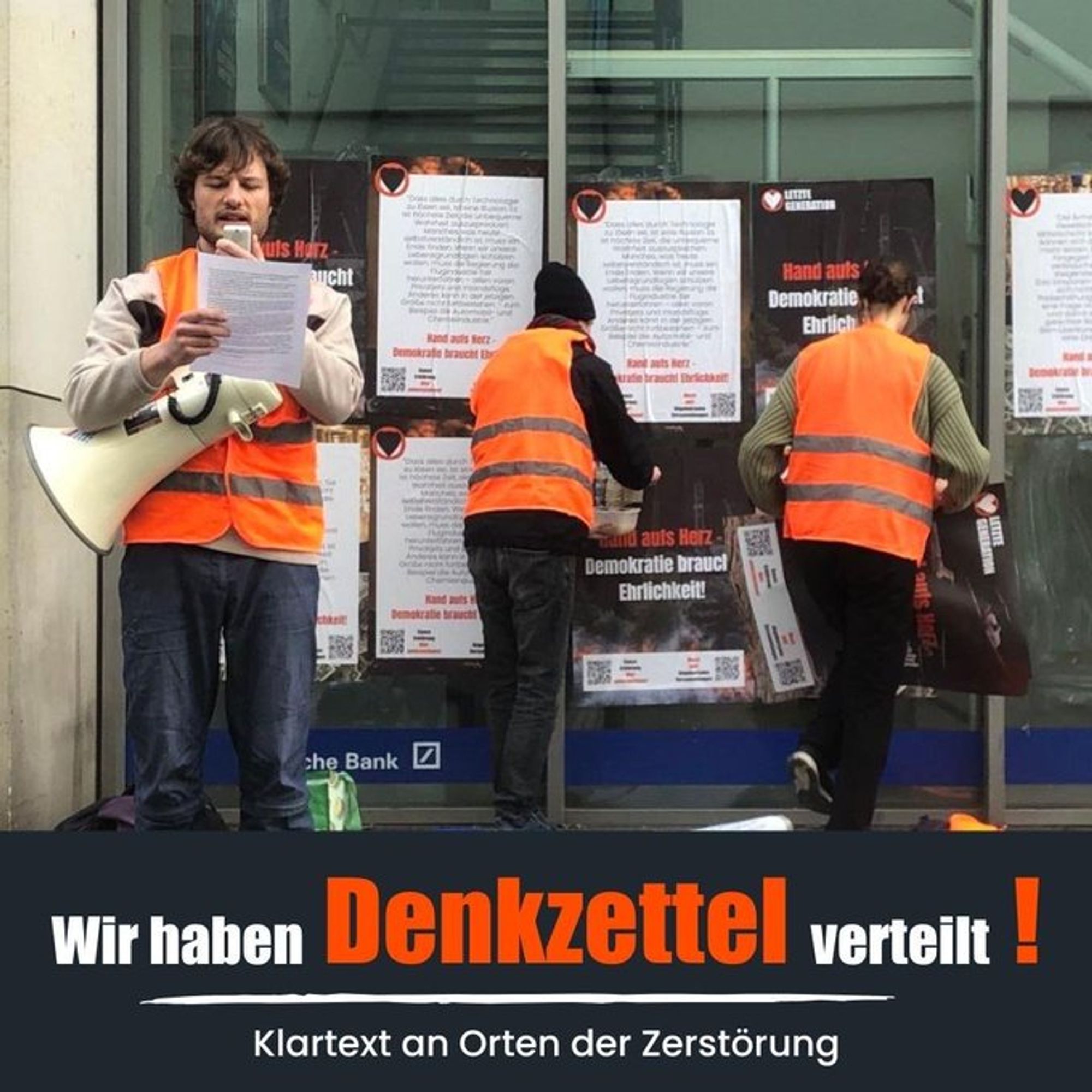 Menschen in orangen Westen kleben Plakate an ein Glasfassade. Davor steht ein Mensch mit Megafon, der eine Rede verliest.
Text: Wir haben Denkzettel verteilt! Klartext an Orten der Zerstörung