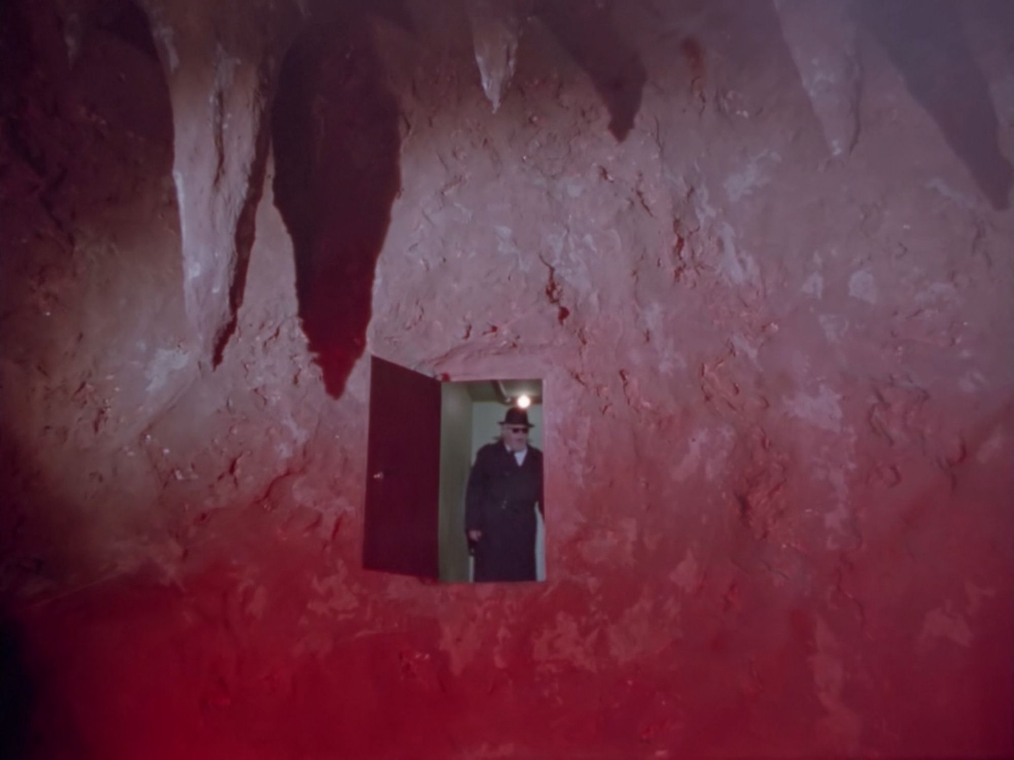 A g-man in a black trenchcoat stands in an open door which is incongruously placed in a vast red cavernous wall