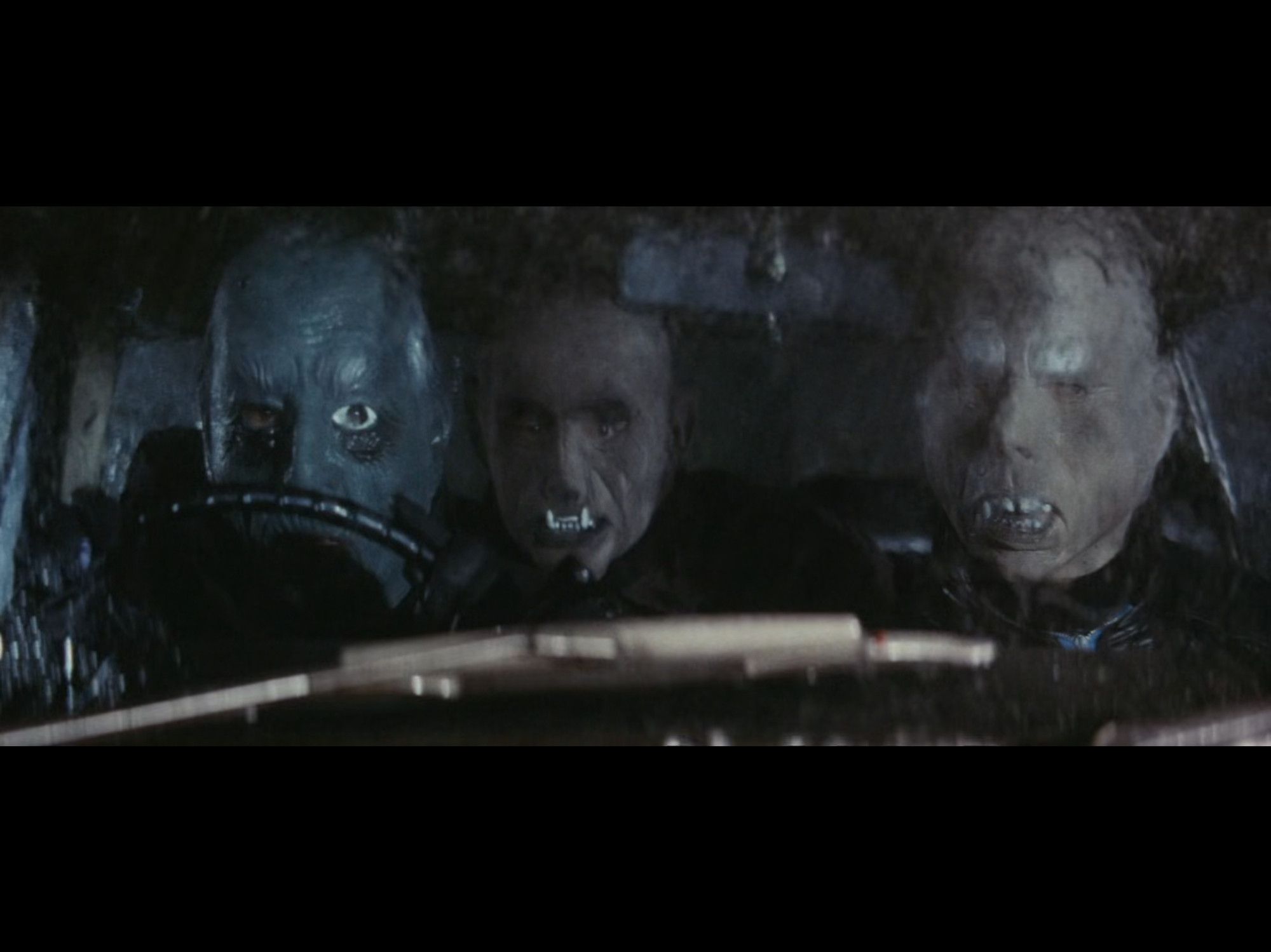 three people in a car as seen through a rainy windshield, all wearing weird rubber monster masks