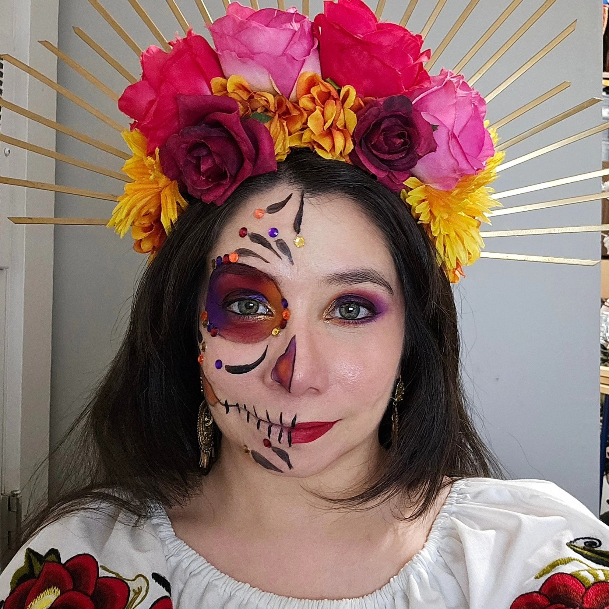 Sophie is wearing a colorful red, pink, and yellow flower headpiece with gold rays. Half of her face is in normal purple, red, and yellow eyeshadow makeup with a red lip. The other half of her face is in a Catrina (decorative skull makeup) using the same colors. She has on a traditional white Mexican top with red embroidery. She has pale skin and long, straight, dark brown hair. The background is a plain white wall.