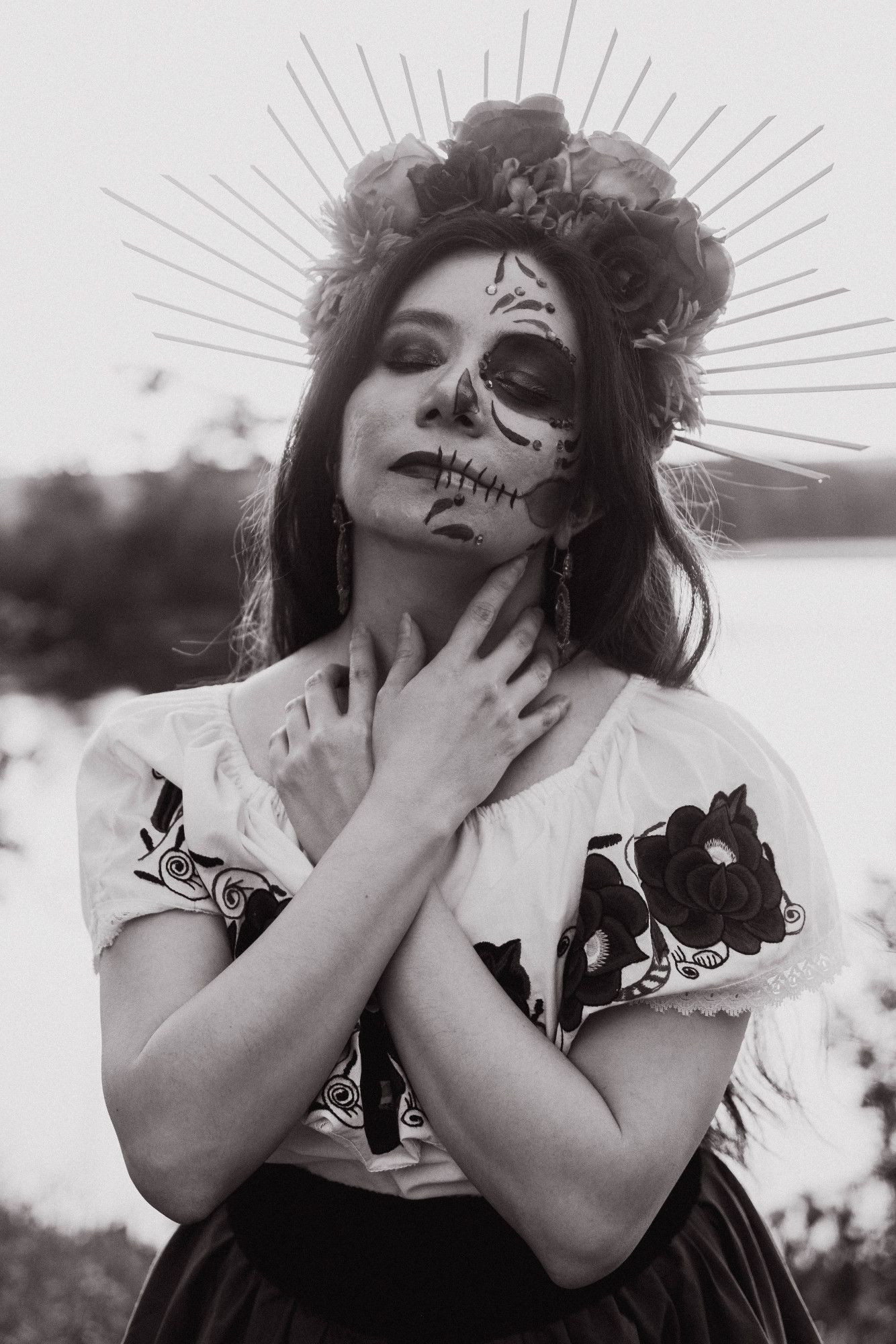 This is a black and white photo of Sophie, who has half of her face in normal makeup, and half of her face in a Catrina (decorative skull makeup). She is wearing a flower headpiece with rays on her long, dark, straight hair. She is wearing earrings, a white traditional Mexican shirt with flower embroidery, a black fabric belt, and a dark skirt. Her hands are draped loosely around her neck and chest.