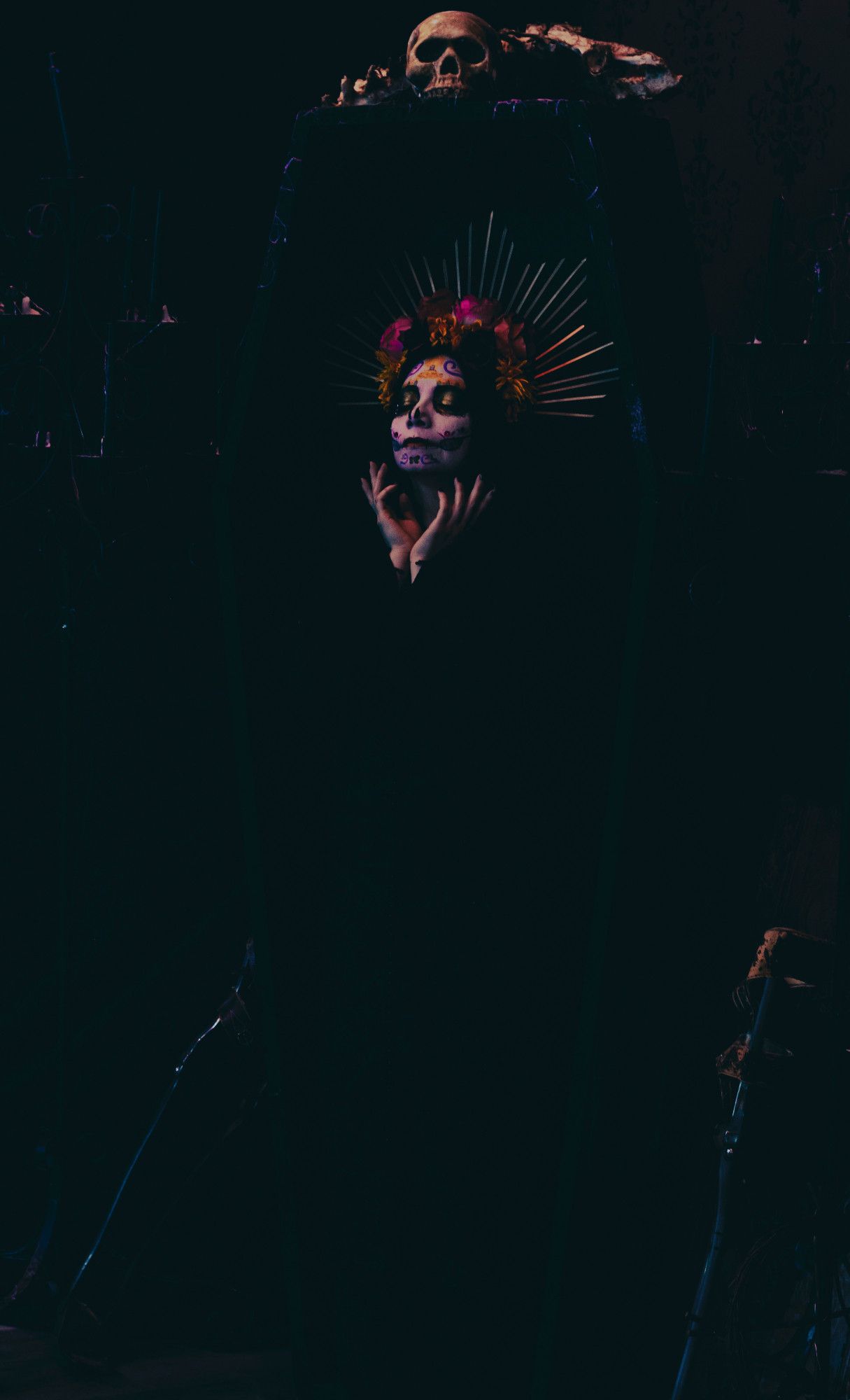 Sophie is wearing black and fading into the black coffin she is standing in. The coffin has a skull and bones on top. Her hands are clawed gently around her face. Her eyes are closed and she is in colorful Catrina makeup (decorative skull makeup). She is wearing a bright rose and marigold flower crown with gold rays.