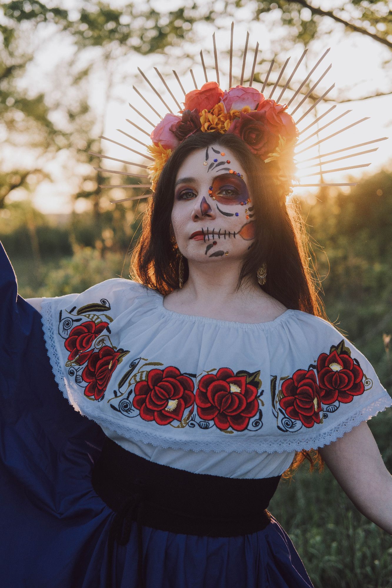 Sophie is looking at the camera with half of her face in normal makeup, and half of her face in a yellow, orange, red, and purple Catrina (decorative skull makeup). She is wearing a colorful flower headpiece with rays on her long, dark, straight brown hair. She is wearing gold earrings, a white traditional Mexican shirt with red flower embroidery, a black fabric belt, and a purple skirt.