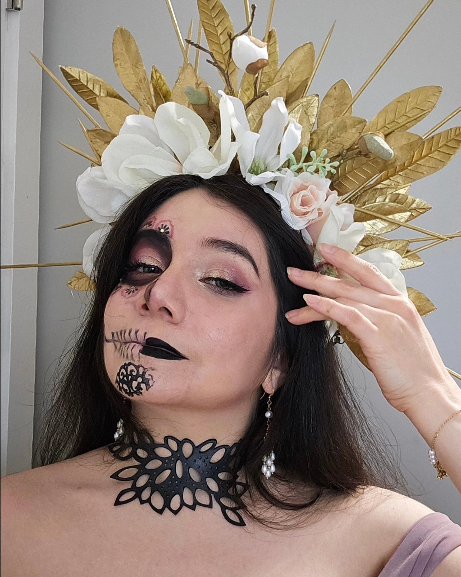 Sophie is wearing a headpiece made of gold leaves, magnolias, and gold spikes. Half of her face is in normal pink and gold eyeshadow makeup with a black lip. The other half of her face is in a Catrina (decorative skull makeup) using the same colors. She has pale pink dress, a black leather choker in a leaf pattern, and dangling pearl earrings. Her skin is pale and her hair is long, straight, and dark brown. he background is a plain white wall.