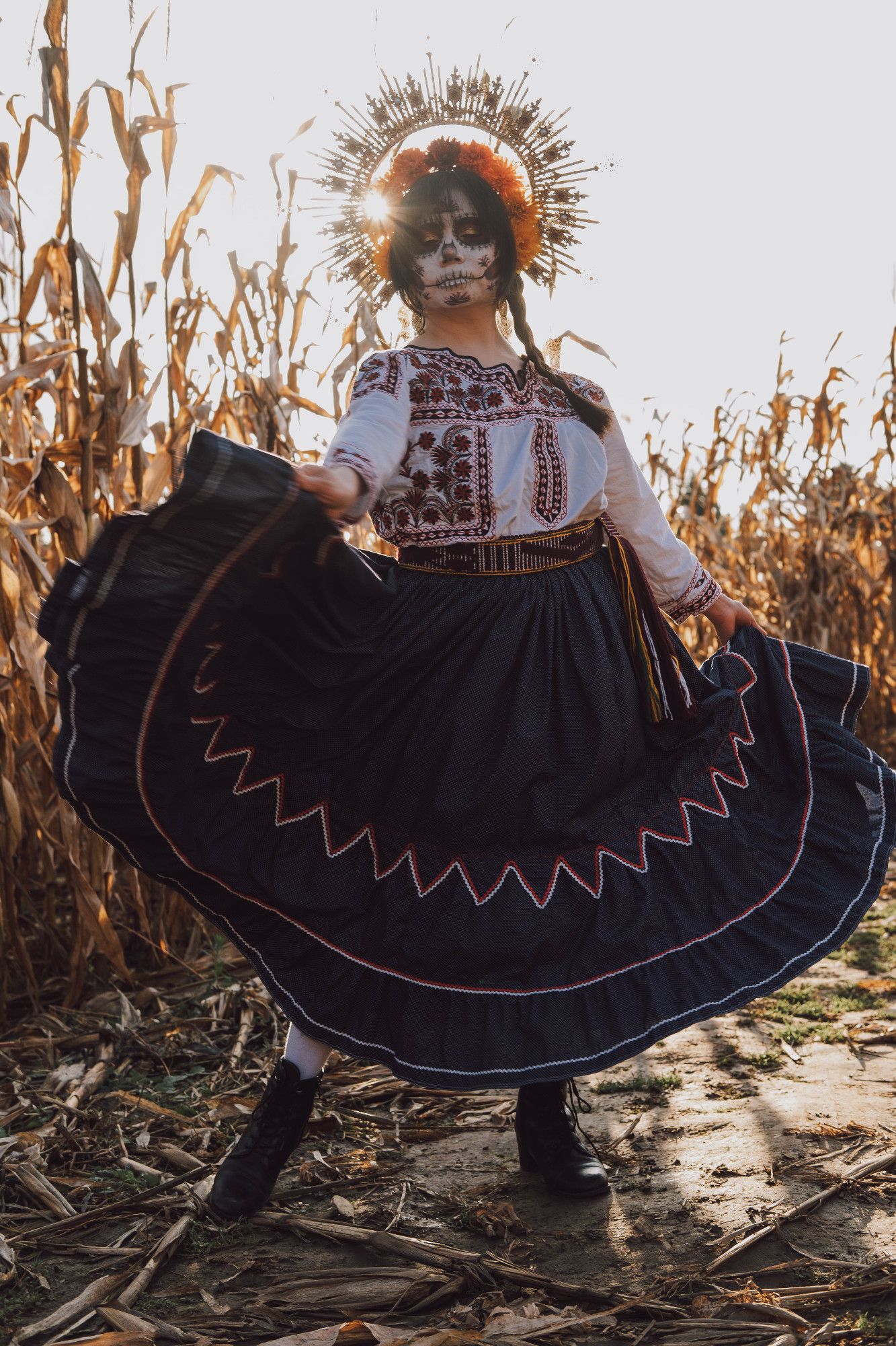 Sophie is standing in a dry cornfield. She is wearing a gold halo filigree headpiece with pearls and red rhinestones; the sun's rays are peeking through the gold There is a halo of red and orange marigolds above her dark hair, which is in two braids. She is wearing gold filigree earrings, a traditional Mexican shirt with red and black flower Mixtec embroidery, a purple and yellow fabric belt, a blue skirt with red and white trim, and black boot with white socks. Her face is in a Catrina (decorative skull makeup) that has patterns that match the shirt embroidery.  The skirt has been swung out with both hands so that the entire diameter of the skirt can be seen flying in the air, the skirt's edge just up above the boots.