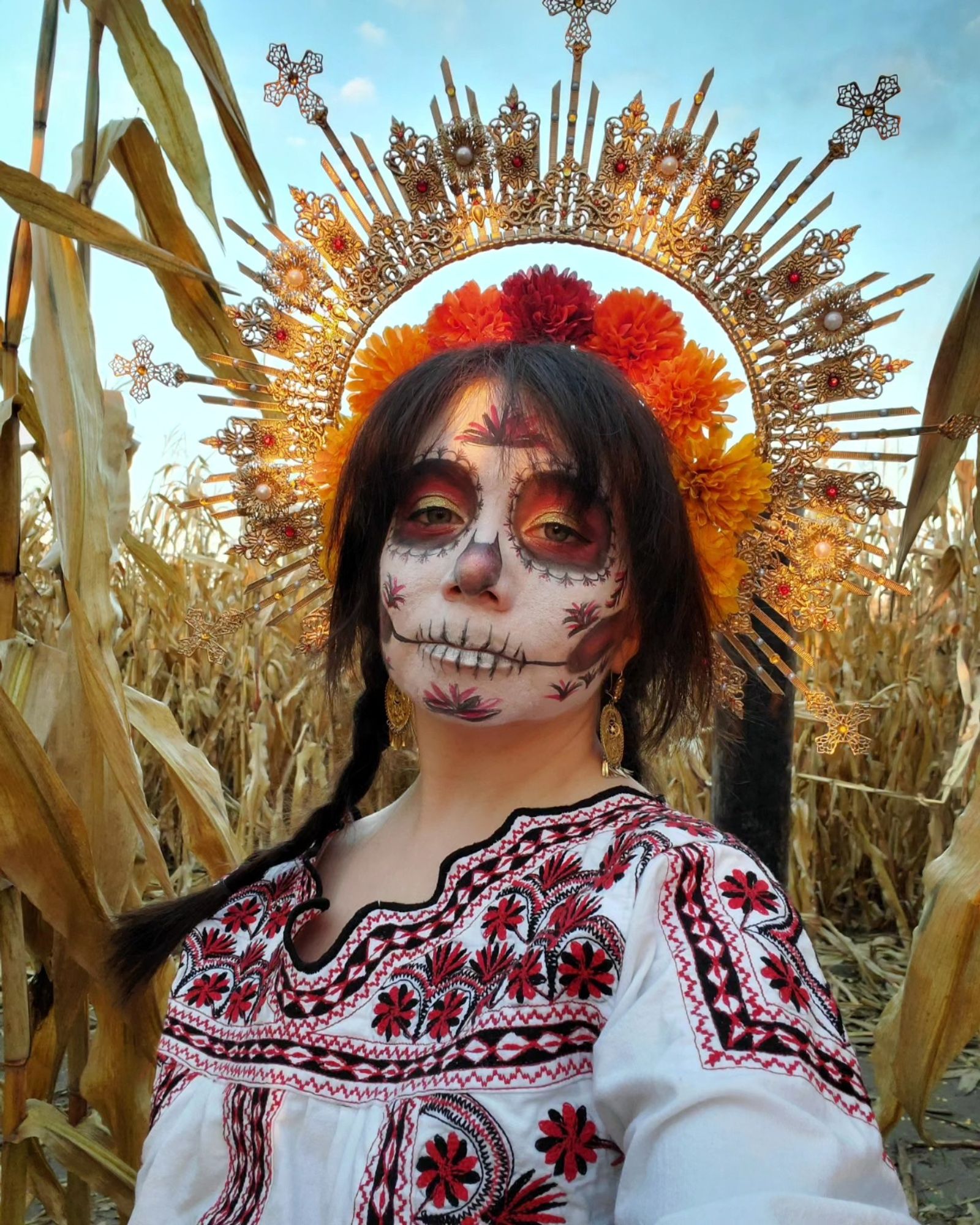 Sophie is outside in the middle of a corn maze with the sun's rays dappled over her. She has on a large gold halo headpiece with filigree, red rhinestones, and pearls. There is a smaller halo of red, orange, and yellow marigolds close to her hair, which is dark brown and in two long braids. She is wearing gold filigree earrings and a white traditional Mexican shirt in black and red Mixtec embroidery. She has pale skin and her face is painted in a white Catrina (decorative skull makeup) with red, gold, and black decorations that match the embroidery.